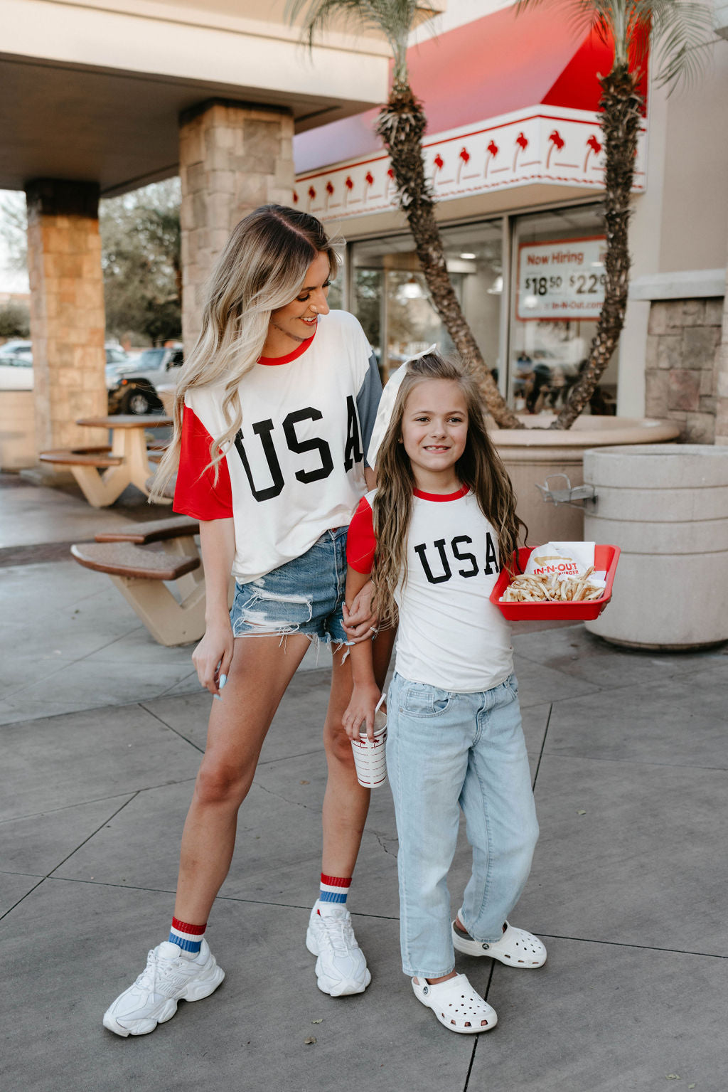A woman and a young girl stand outdoors near picnic tables, both wearing matching forever french baby "Children's Bamboo T-Shirt | USA, Land That I Love" with red sleeves—perfect 4th of July clothes. The woman has a drink, and the girl holds a tray of food and a shake. They smile and look down, likely engaging with each other.