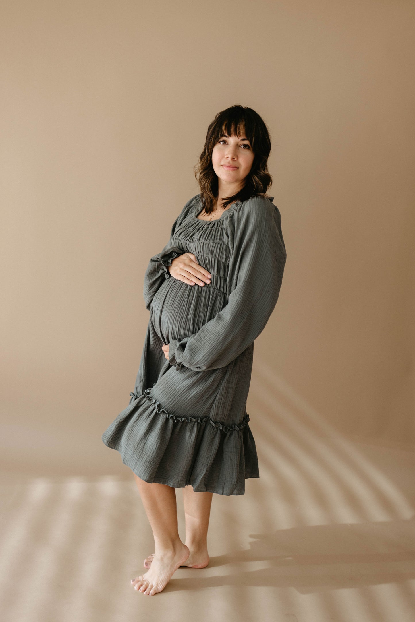 A pregnant woman with shoulder-length brown hair stands barefoot on a beige floor, gently cradling her belly and smiling in a loose, long-sleeved gray Women's Dress | Meadow by forever french baby.