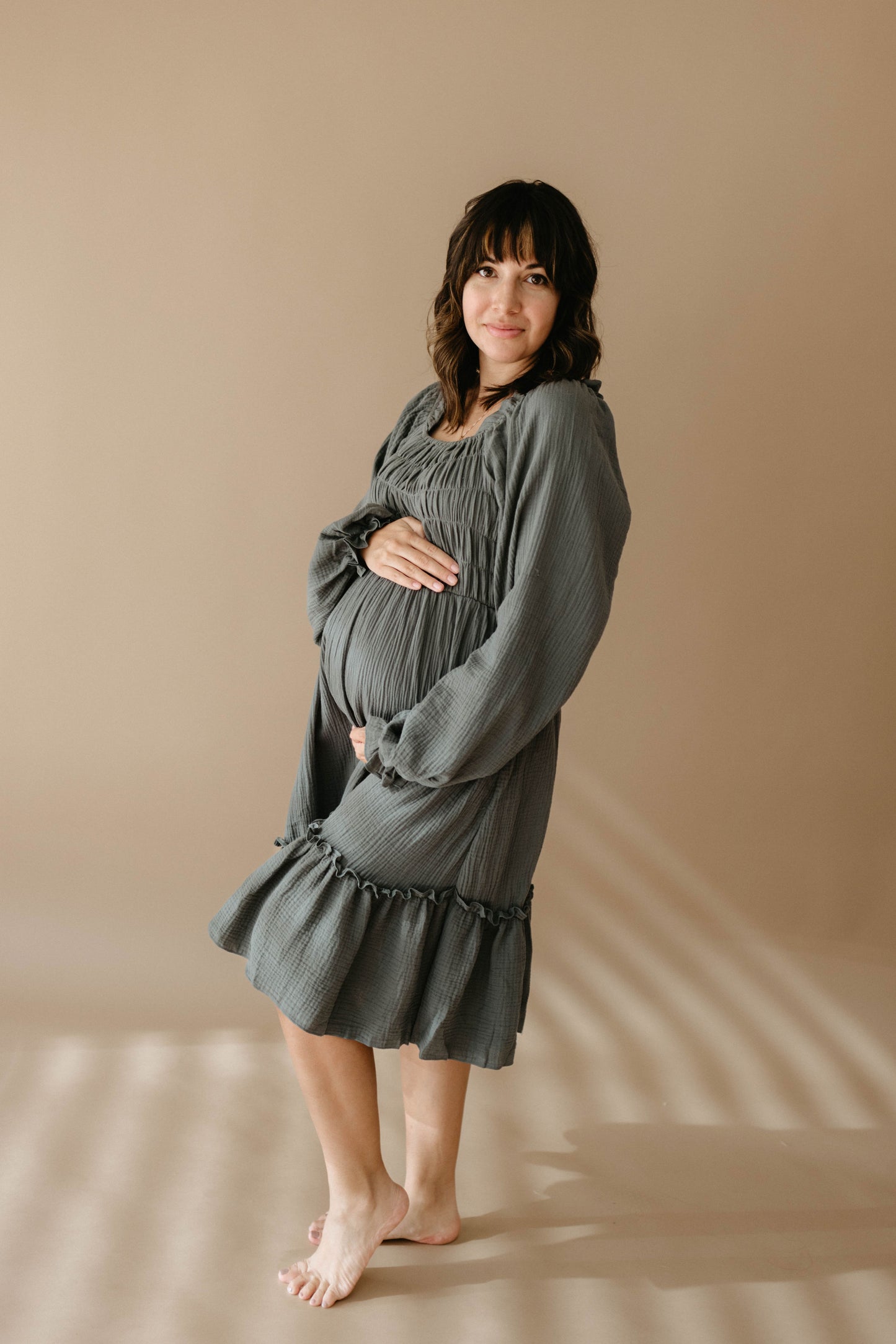 A pregnant woman with shoulder-length brown hair stands barefoot on a beige floor, smiling softly at the camera while wearing the loose-fitting, long-sleeved Meadow dress by forever french baby and gently cradling her belly.
