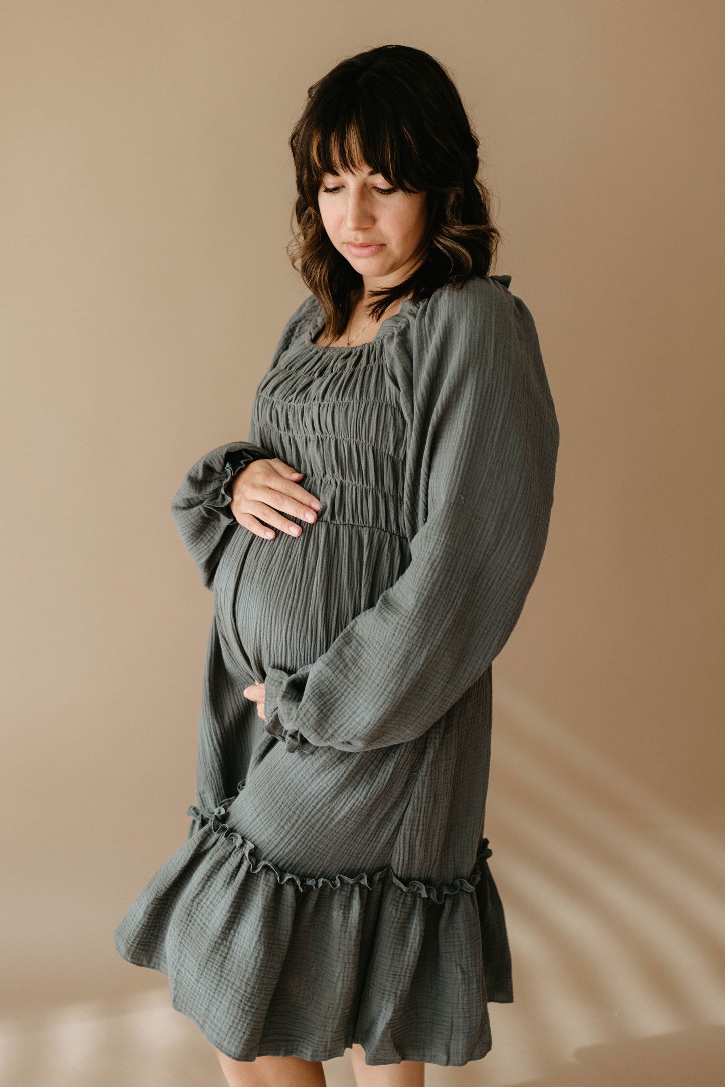 A pregnant woman cradles her baby bump and looks down softly, wearing the Meadow women's dress by forever french baby, standing against a plain beige background.