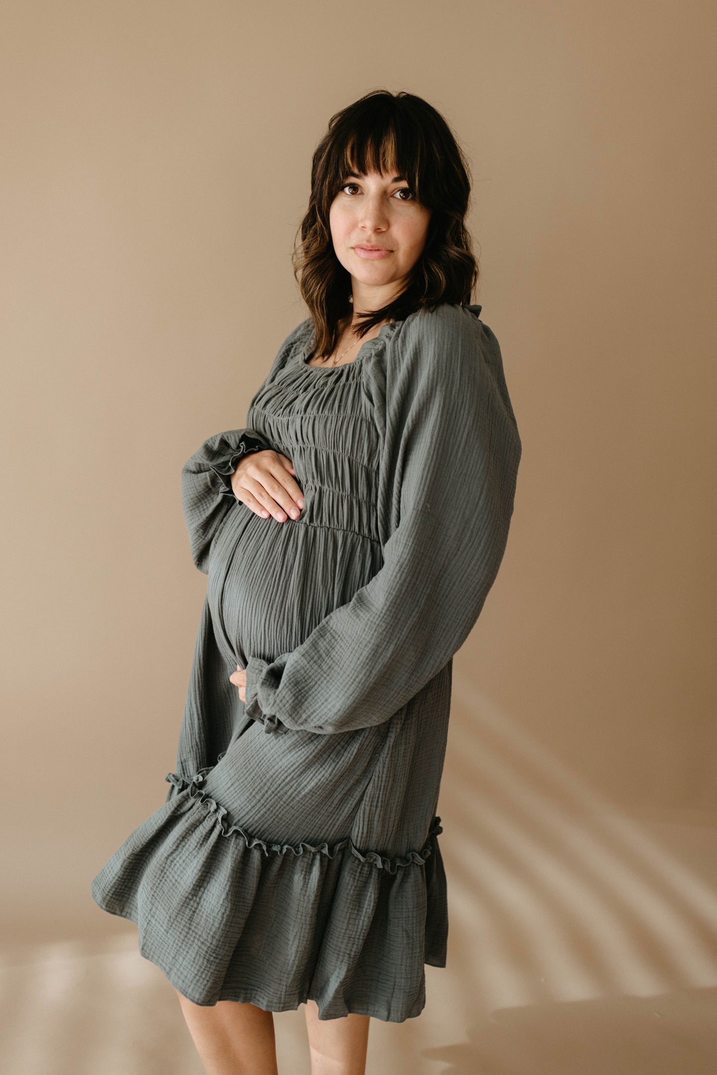 A pregnant woman with medium-length dark hair wears the Meadow women's dress from forever french baby, standing against a beige background and gently holding her belly while looking at the camera.