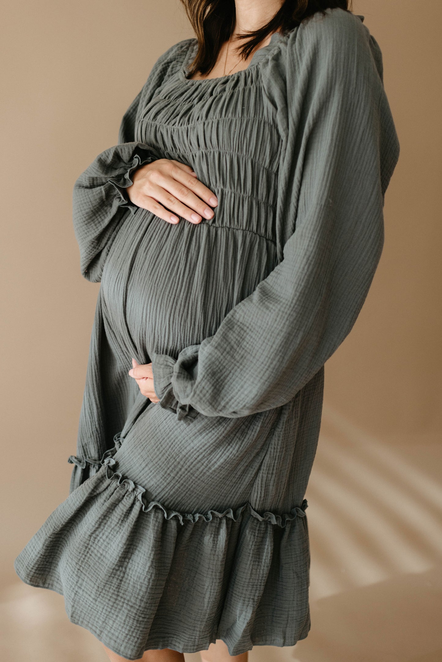 A pregnant woman, face unseen, gently cradles her belly in a loose, textured Women's Dress | Meadow by forever french baby against a soft beige background.