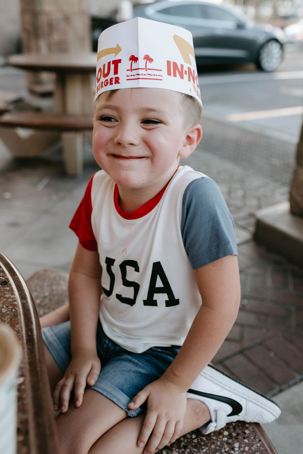 A young child sitting outdoors, smiling, and wearing a paper hat from In-N-Out Burger. The child is dressed in eco-friendly 4th of July clothes from forever french baby, featuring a red, white, and blue Children's Bamboo T-Shirt with the letters "USA" on it and denim shorts. The background shows a casual outdoor seating area.