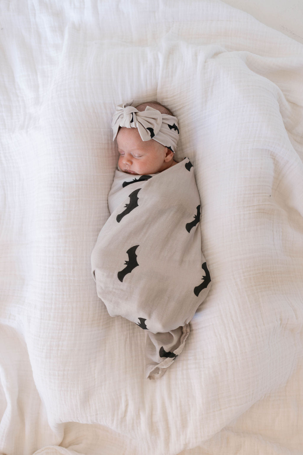 A baby is snugly swaddled in the Muslin Swaddle | It's Bats! from forever french baby, made of soft muslin and featuring a charming beige bat print. The baby wears a coordinating beige headband tied into a bow and rests peacefully on a soft, white quilted surface.