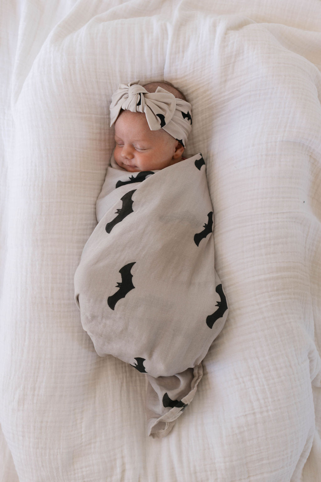 A sleeping baby is snugly wrapped in a **Muslin Swaddle | It's Bats! by forever french baby**. The baby lies on a plush, white surface, wearing a matching beige headband with a bow. The serene scene exudes calmness and warmth.