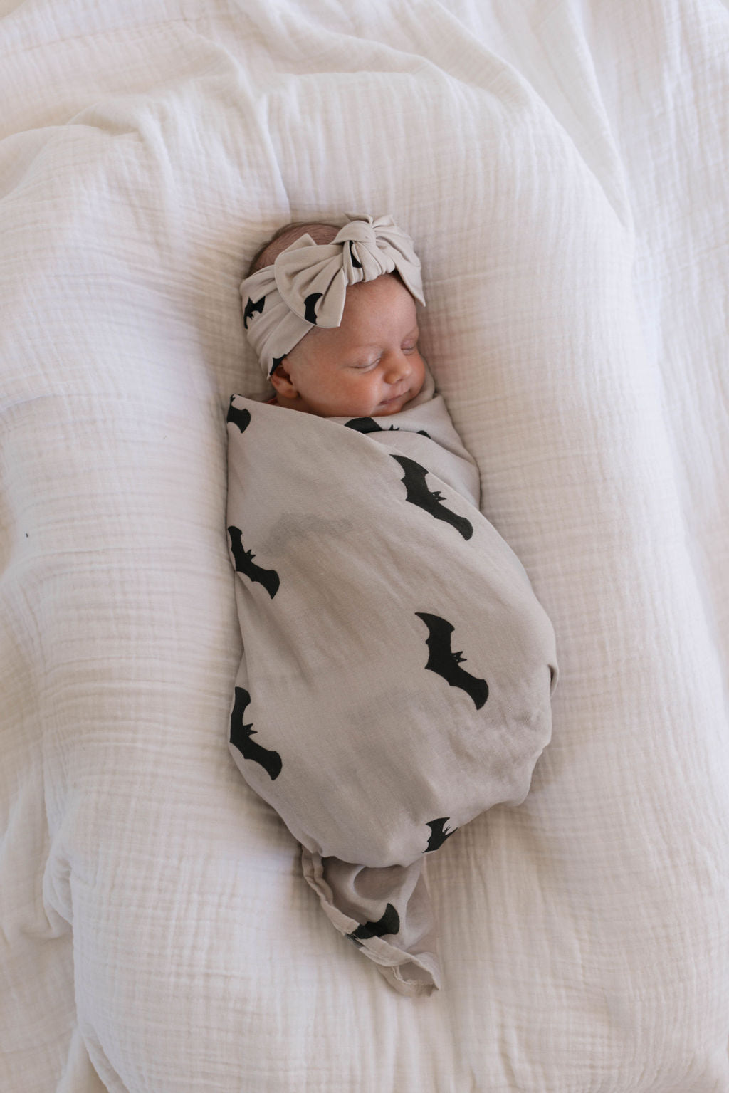A sleeping baby is cozily wrapped in the **forever french baby Muslin Swaddle | It's Bats!**, complemented by a matching headband with a bow. The baby is laying on a soft, white, textured blanket.