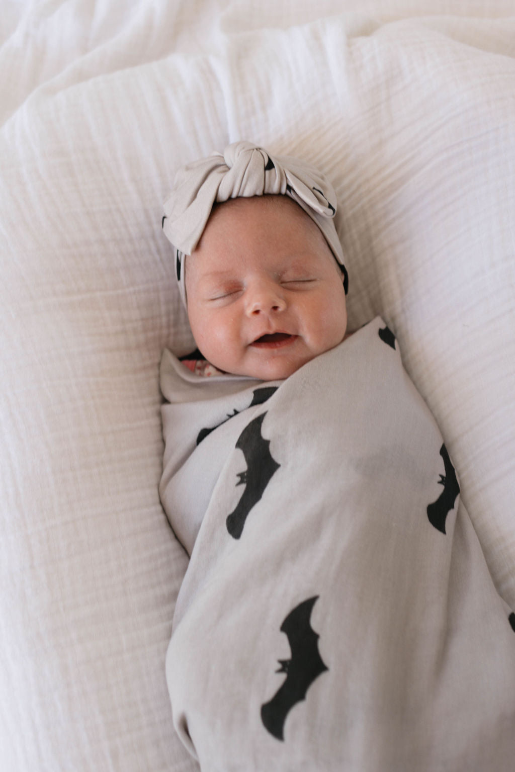 A smiling baby is swaddled in a Muslin Swaddle | It's Bats! from forever french baby. The baby is lying on a soft, white surface and is wearing a matching light gray headband with a bow.