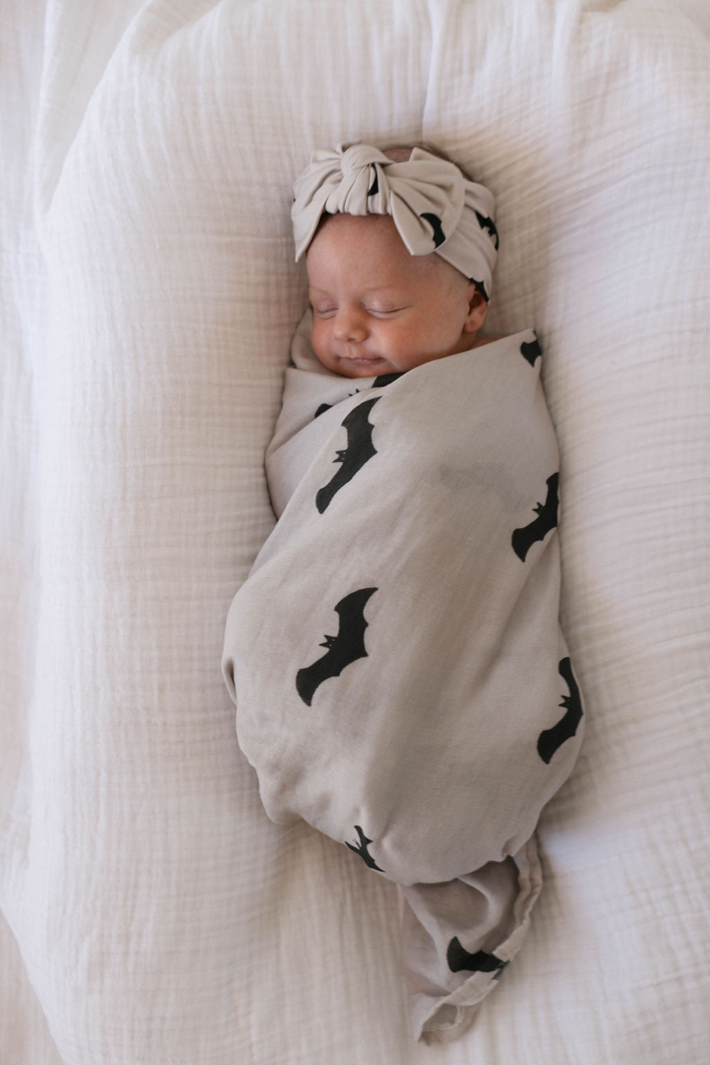 A newborn baby is swaddled in the forever french baby Muslin Swaddle featuring the adorable It's Bats! print. The baby is wearing a matching beige headband with a bow and is peacefully sleeping on a white cushioned surface.