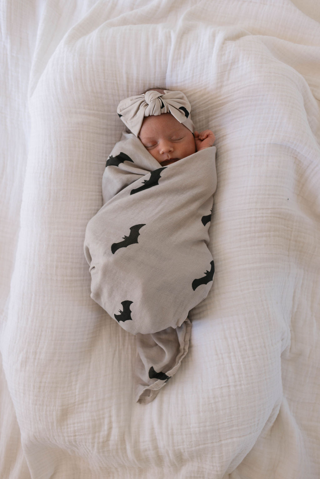 A newborn baby is swaddled in the forever french baby's Muslin Swaddle from the It's Bats! collection, featuring a light gray background with black bat silhouettes. The baby is wearing a coordinating gray headband with a bow and is peacefully sleeping on a white textured blanket.