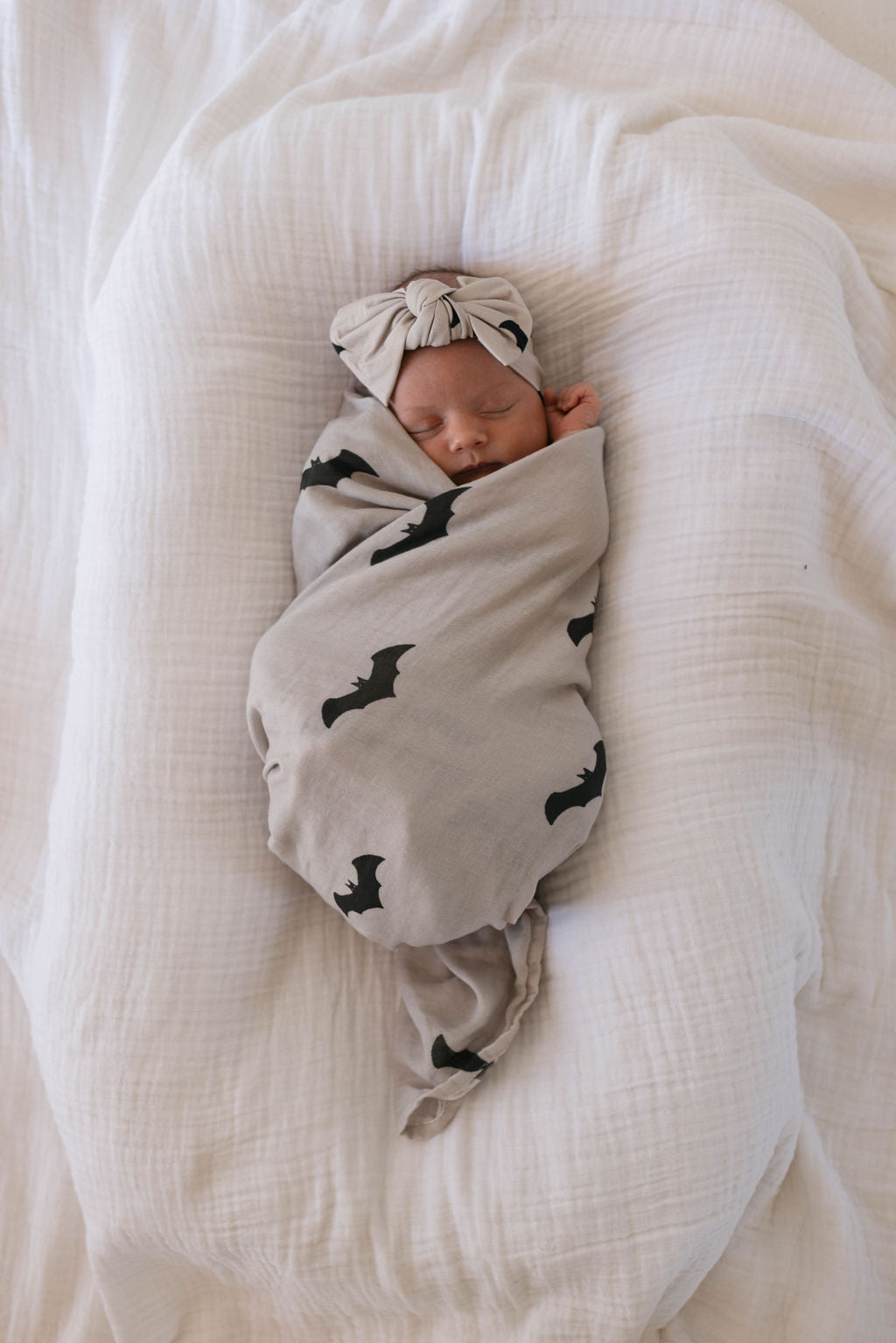 A sleeping baby is swaddled in a soft Muslin Swaddle | It's Bats! print blanket by forever french baby, lying on a white bed. The baby is wearing a matching headband bow, and the scene looks peaceful and cozy.