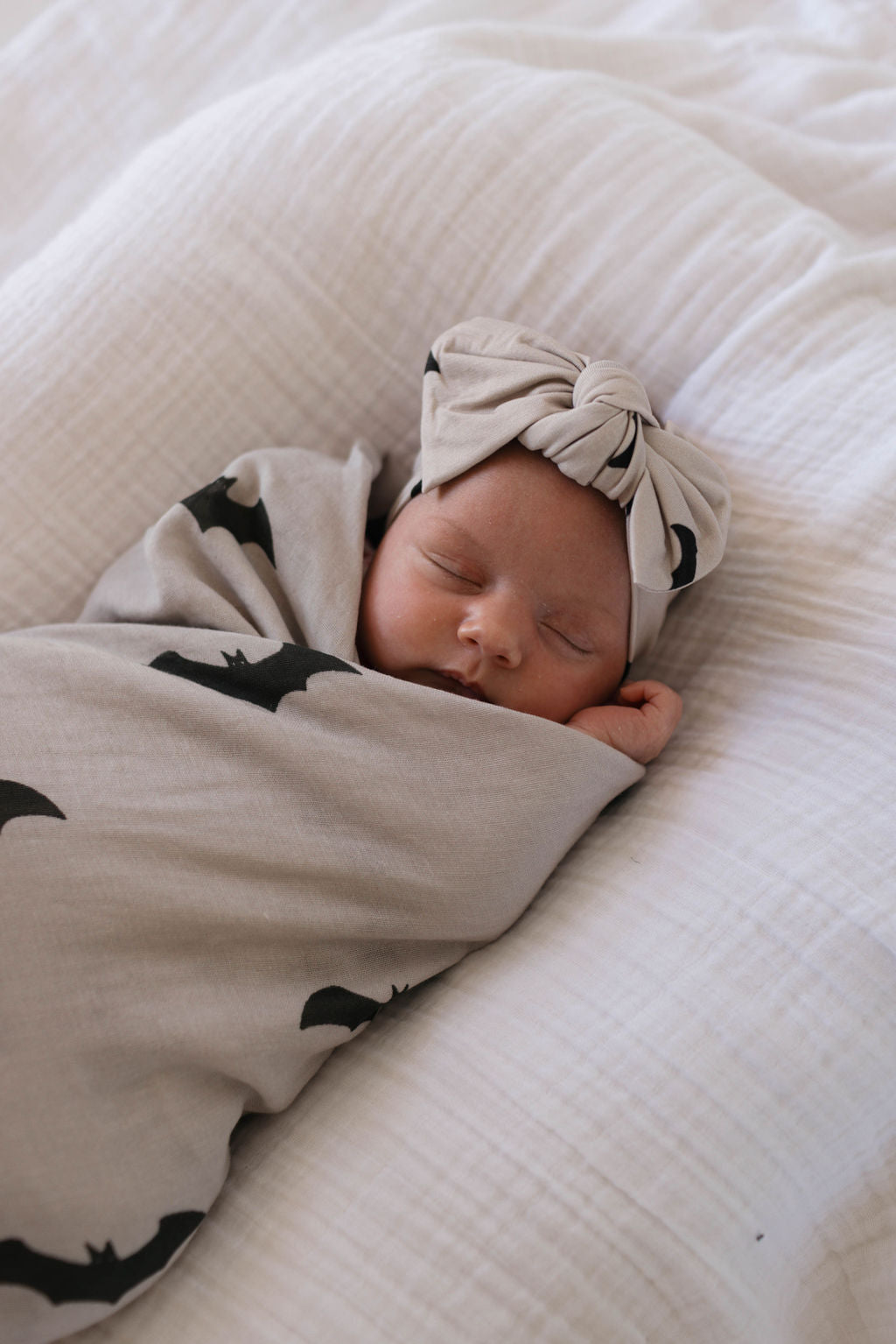 A newborn baby, peacefully sleeping with eyes closed and arms tucked in, is warmly wrapped in a soft Muslin Swaddle from forever french baby featuring the charming It's Bats! print. The baby lies on a white, textured blanket and wears a matching beige headband with a bow.