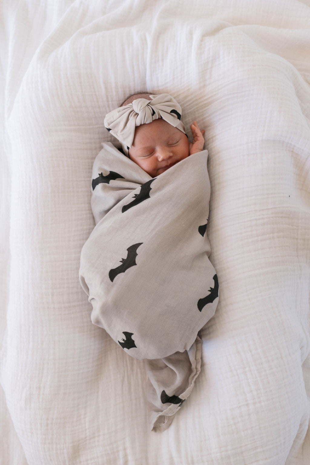 A newborn baby is sleeping peacefully, wrapped in a beige muslin swaddle adorned with black bat designs from the It's Bats! collection by forever french baby. The baby also wears a matching headband, lying on a soft, white blanket.