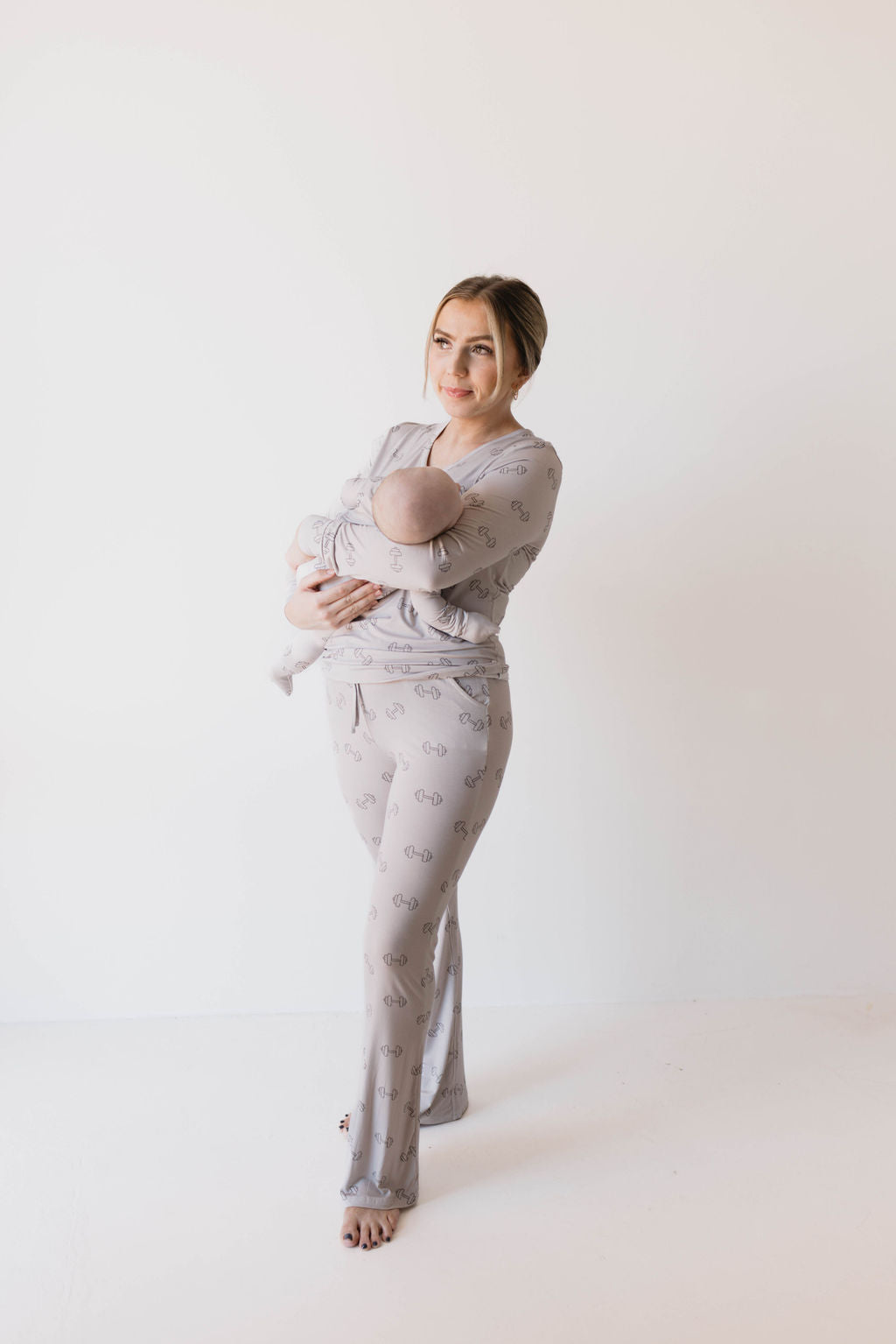 A woman, standing against a plain white background, is wearing the Women's Bamboo Pajamas | Arm Day from forever french baby, a matching light gray pajama set with a subtle pattern. These pajamas feature breastfeeding accessibility. She is cradling a baby in her arms, and both look content and relaxed.