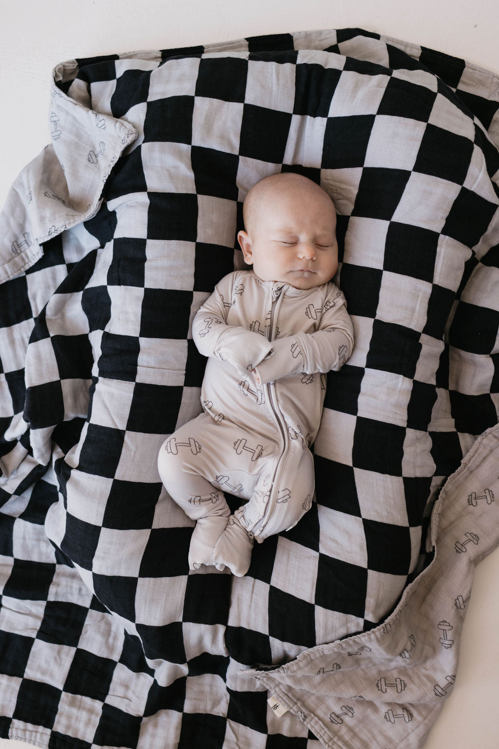 A baby wearing a light-colored onesie with subtle patterns sleeps peacefully on a Muslin Quilt from forever french baby, featuring the Arm Day + Black & Grey Checkerboard design. The baby is swaddled, and the slightly wrinkled blanket adds to the cozy atmosphere.