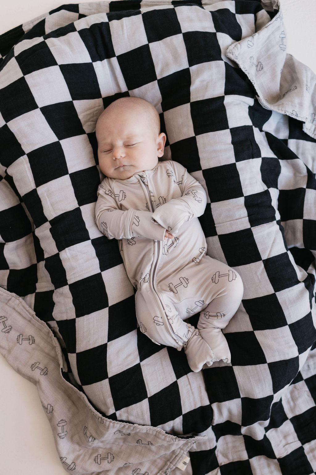 A baby sleeps peacefully on a black and grey checkerboard blanket. The baby is wearing a light grey onesie with a simple pattern, their arms crossed over their chest. The Muslin Quilt | Arm Day + Black & Grey Checkerboard from Forever French Baby, spread out beneath them, provides a cozy and comfortable surface.