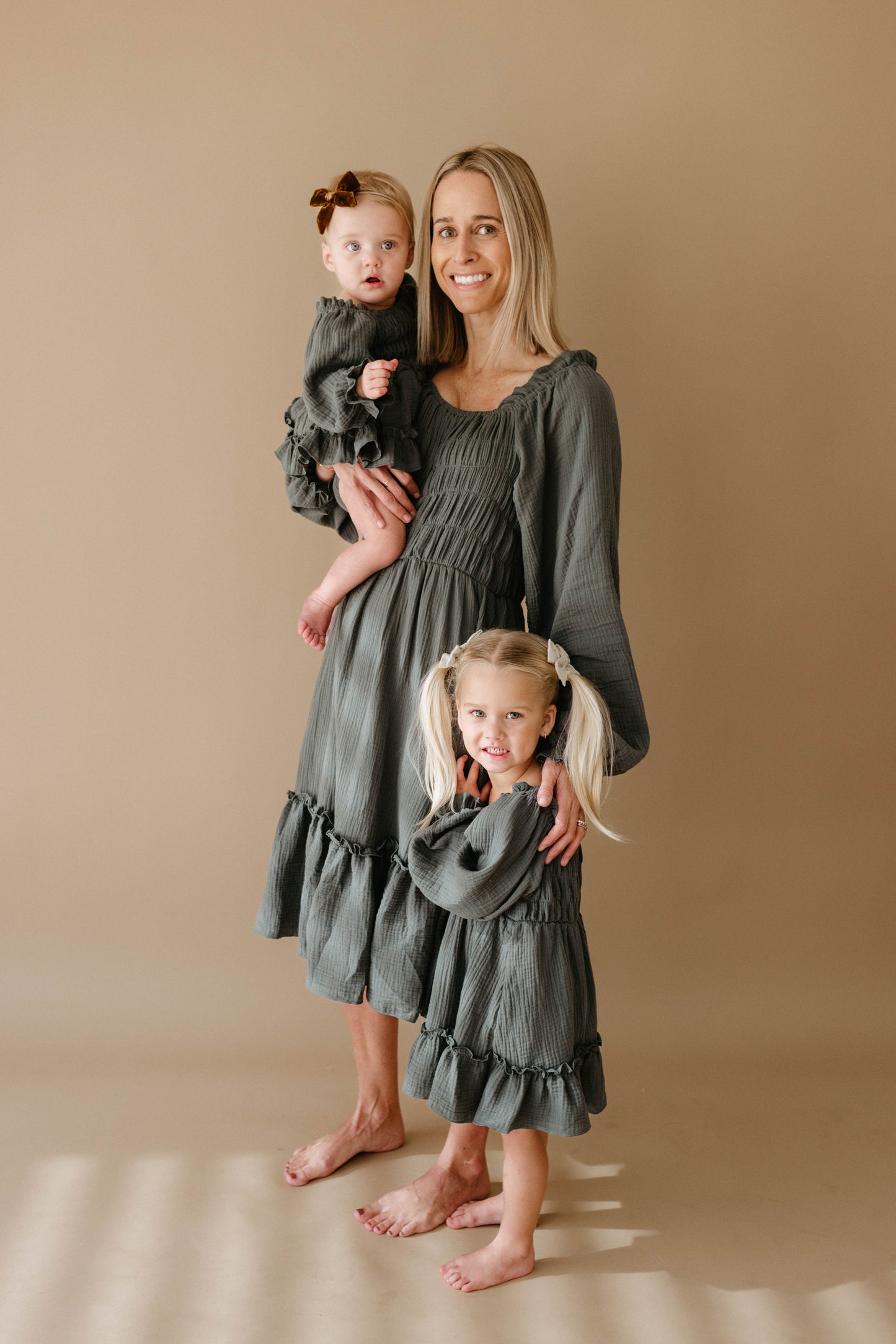 A woman stands barefoot holding a young girl while another toddler stands beside her; all wear matching forever french baby Meadow dresses, featuring long ruffles in gray-green, posed against a plain beige background.