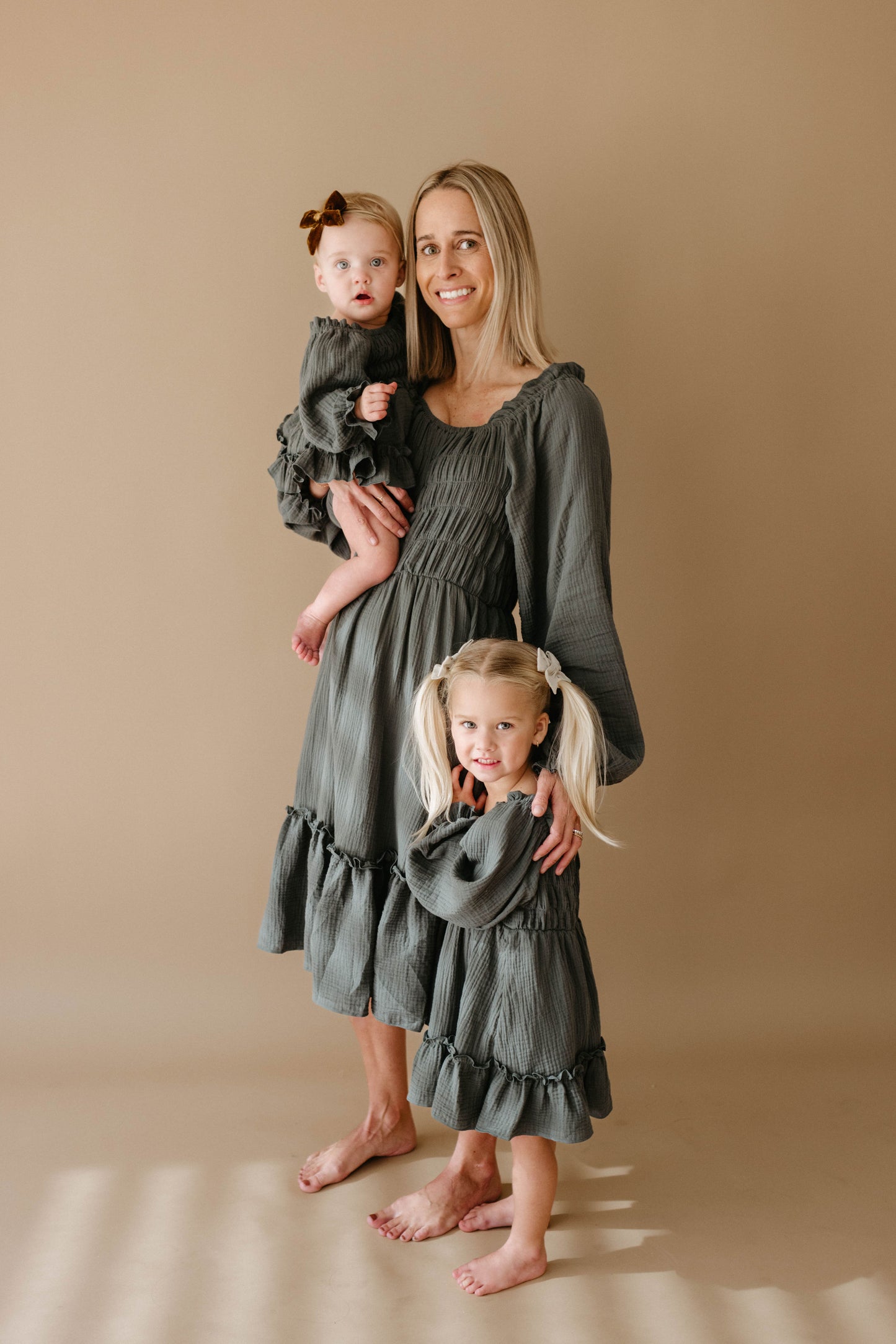 A woman, a toddler with a brown bow, and a blonde girl in pigtails stand barefoot on beige, all wearing matching forever french baby Meadow long-sleeve ruffled olive-green dresses.
