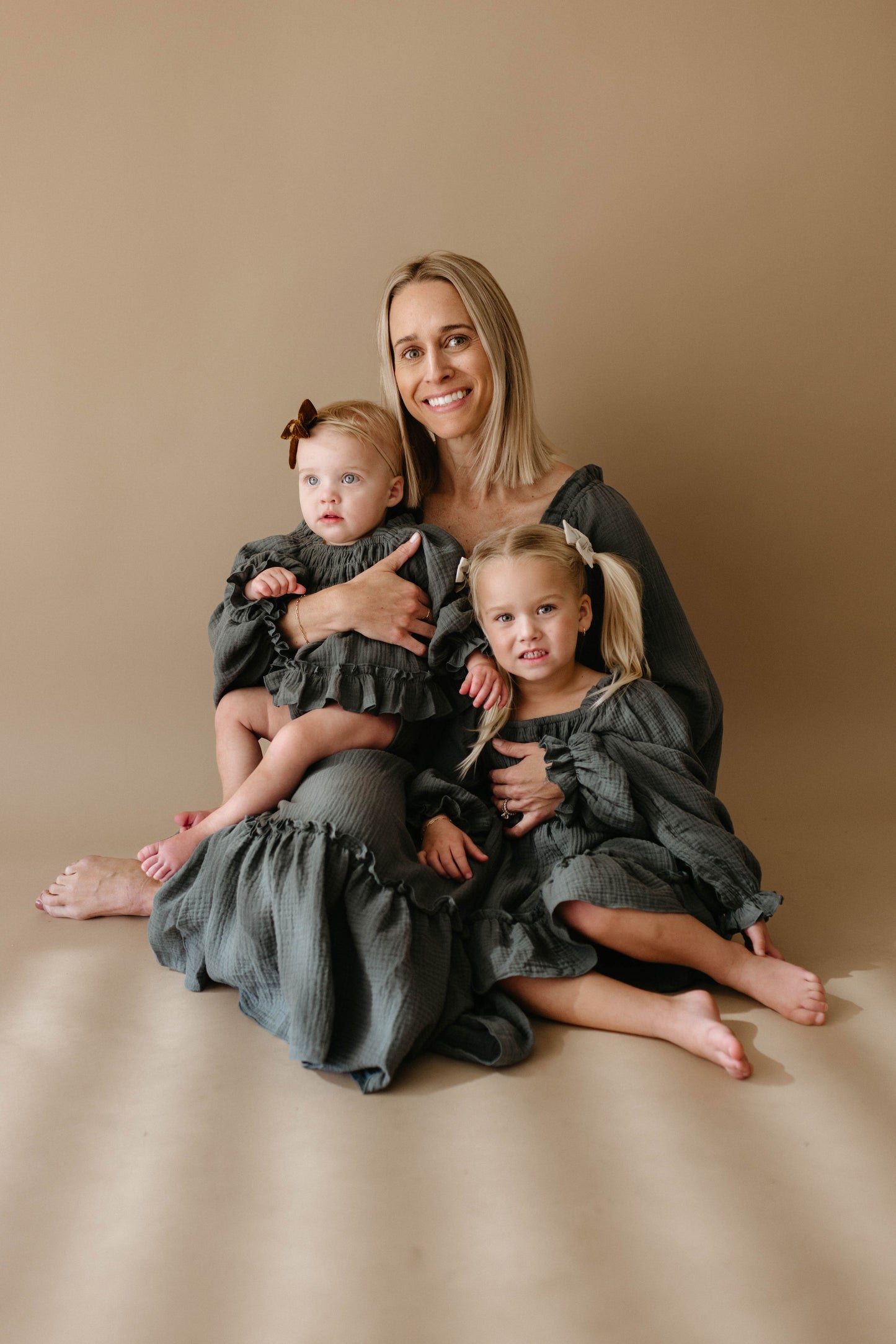 A woman sits barefoot on the floor, smiling and holding two young girls dressed in matching forever french baby Child's Dress | Meadow outfits. Against a beige backdrop, one child sits in her lap while the other is by her side.