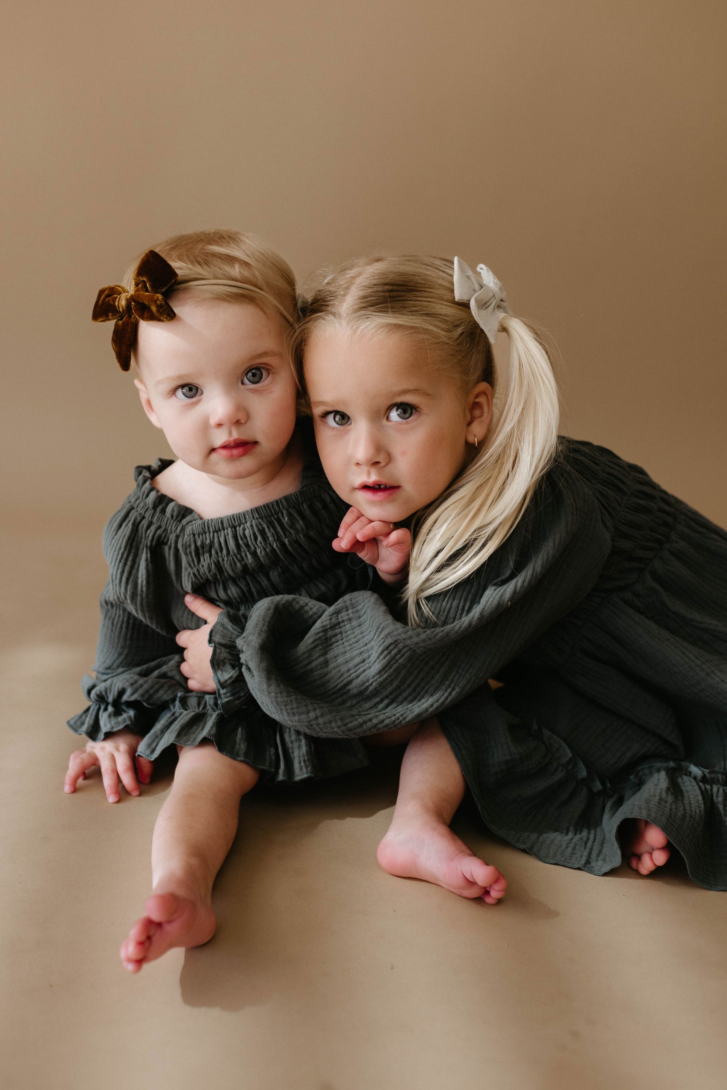 Two young girls sit on the floor against a beige background. Both wear matching "Meadow" dresses by forever french baby; the older hugs the younger, who has short blonde hair and a brown bow.