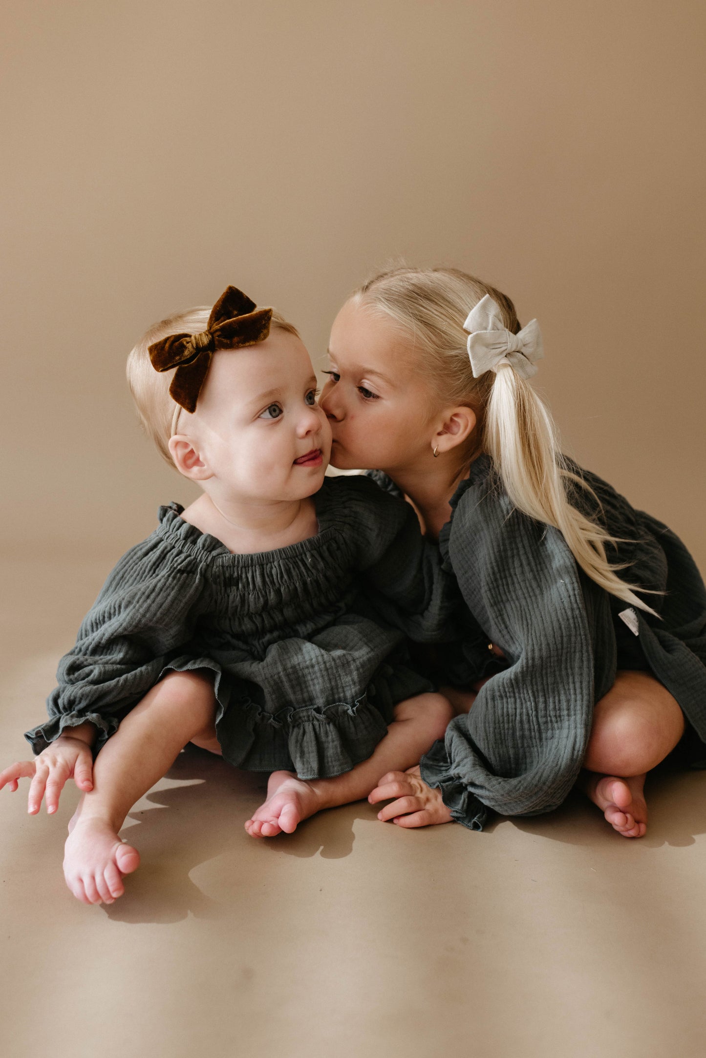 A young girl with a white bow kisses a baby with a brown bow, both barefoot and wearing matching forever french baby's Meadow Child's Dress in dark green, seated against a light brown background.