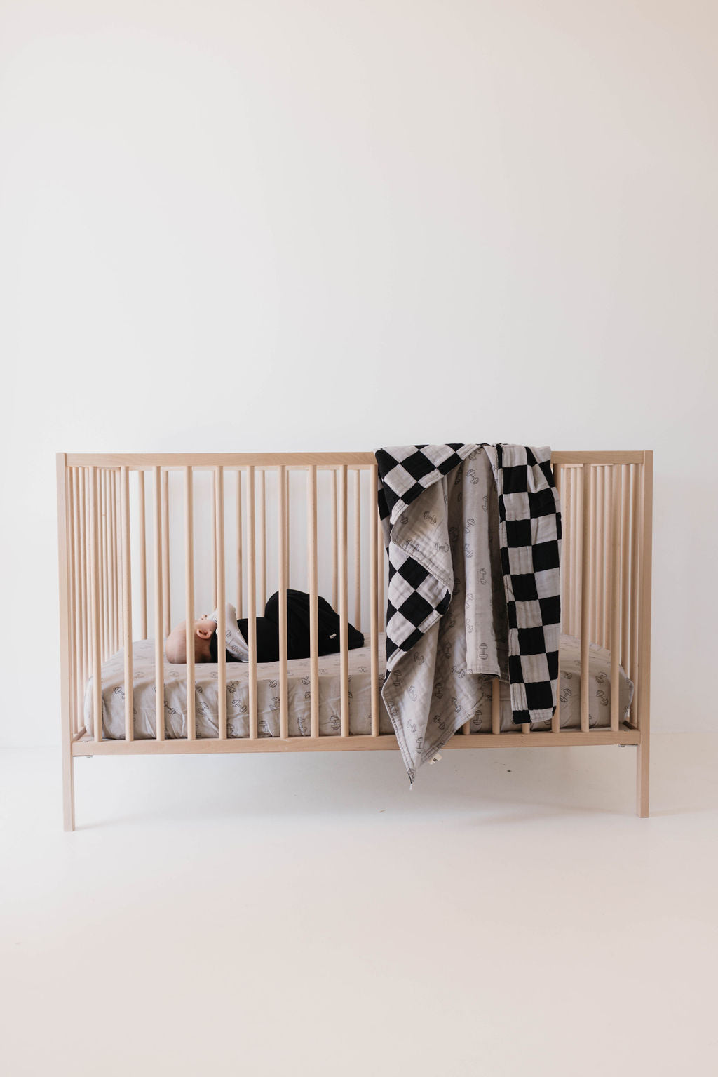 A baby lies on their back in a wooden crib with vertical slats, covered with white bedding. Draped over the side of the crib is a Muslin Quilt in Arm Day + Black & Grey Checkerboard by forever french baby. The background features a plain, light-colored wall.