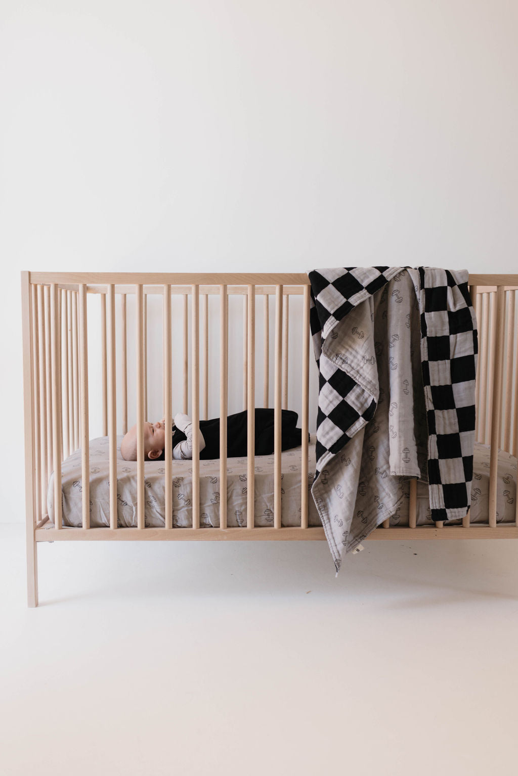 A baby lies on their back in a wooden crib with light gray bedding. Draped over the side of the crib is Forever French Baby's Muslin Quilt in their Arm Day + Black & Grey Checkerboard design. The minimalist room features white walls and floors, while the baby is dressed in dark clothing.