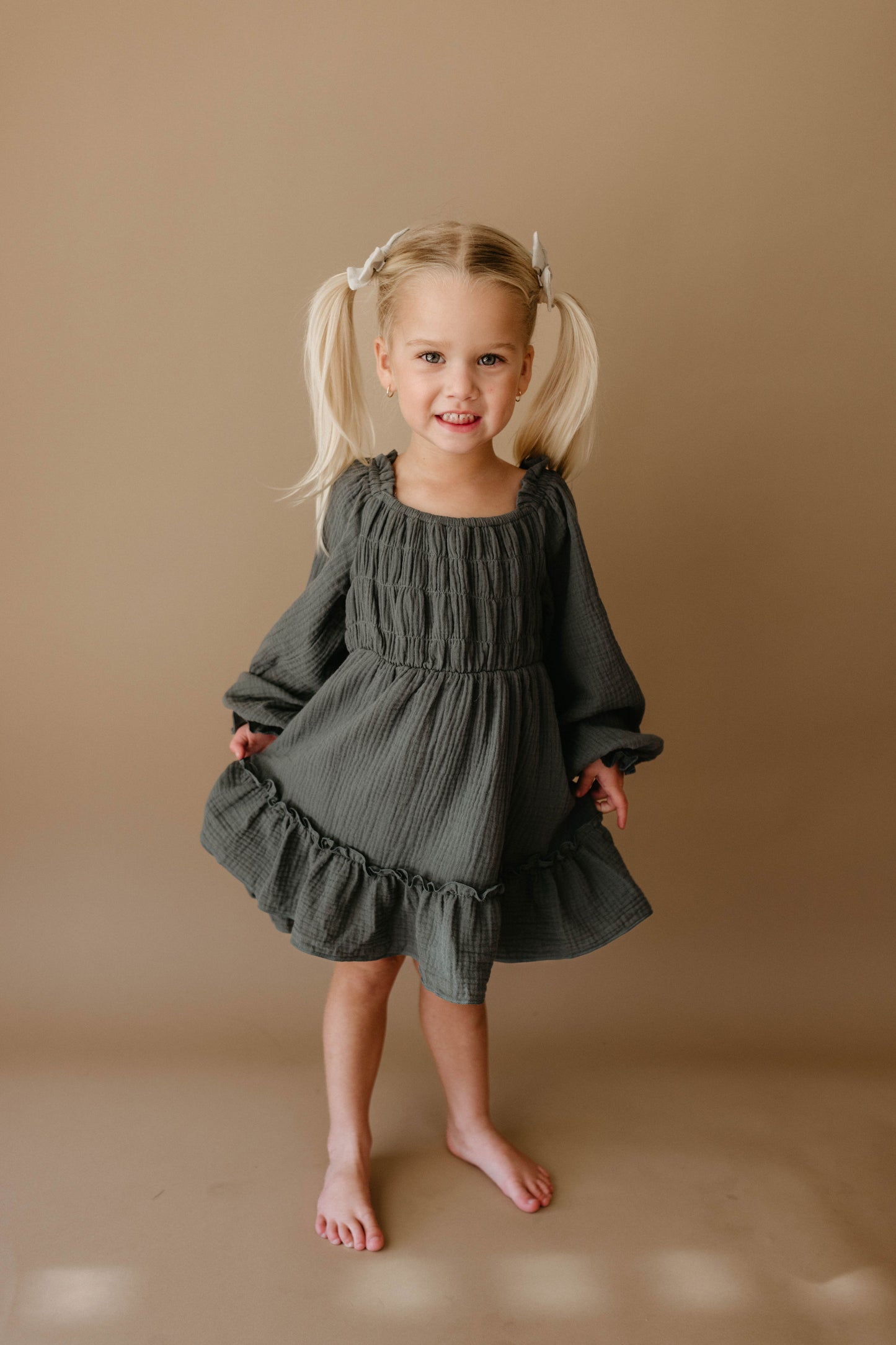 A young girl with blonde pigtails, barefoot and smiling, stands against a beige background wearing the forever french baby Child's Dress | Meadow in dark green, gently holding the sides of her long-sleeved dress.