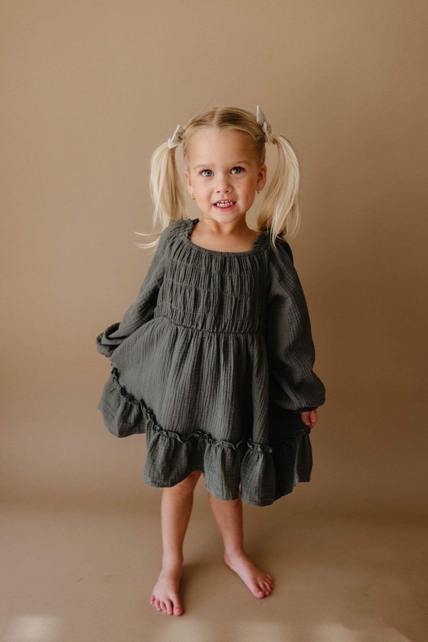 A smiling young girl with blonde pigtails wears the Meadow Child's Dress from forever french baby—a dark green, long-sleeved, ruffled dress—standing barefoot against a neutral background.