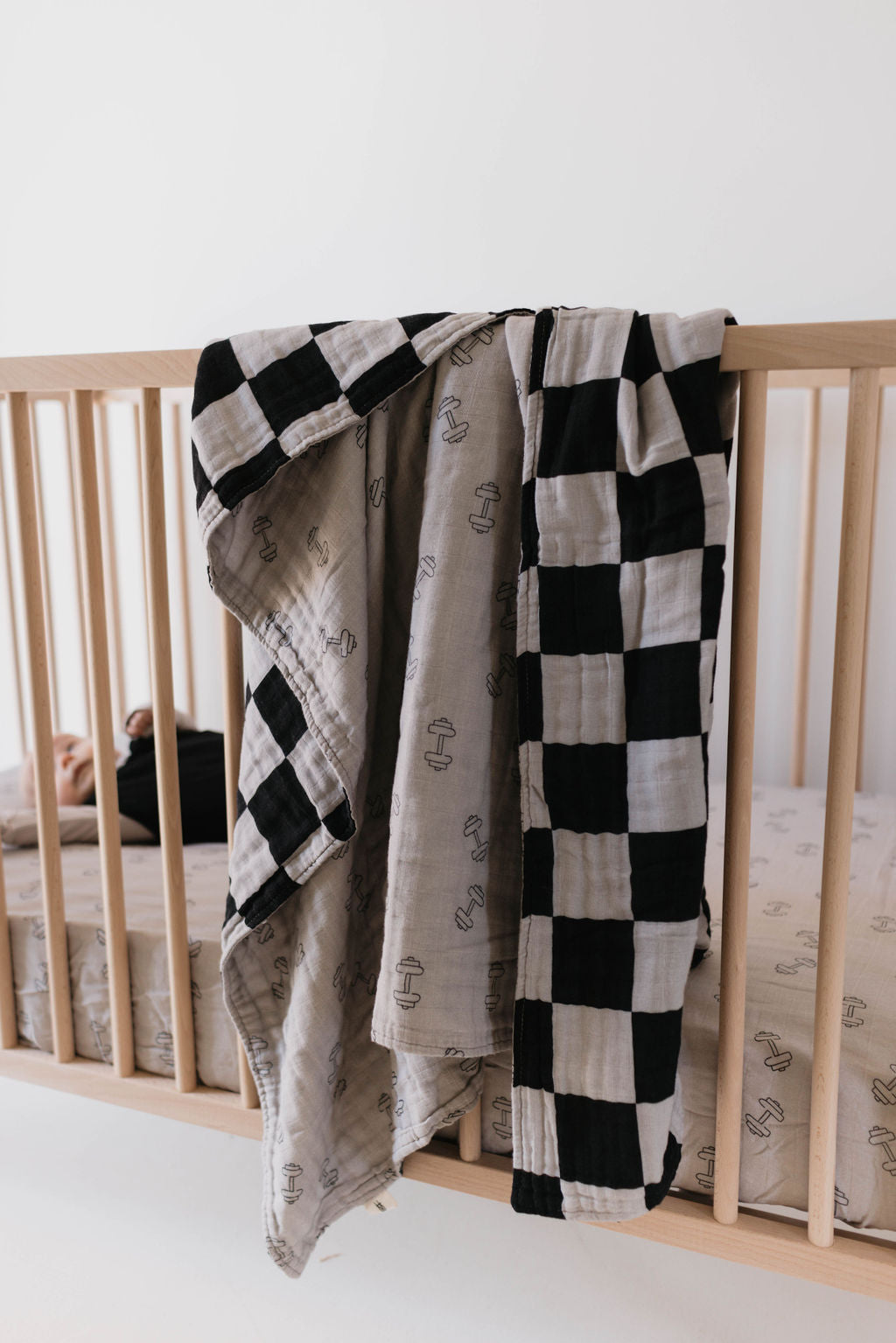 A wooden crib adorned with light-colored bedding features a Muslin Quilt in Arm Day + Black & Grey Checkerboard draped over one side. A partially visible baby, with just a bit of head and arm showing, lies inside the crib. The room's minimalist decor elegantly reflects the sophisticated style of forever french baby designs.