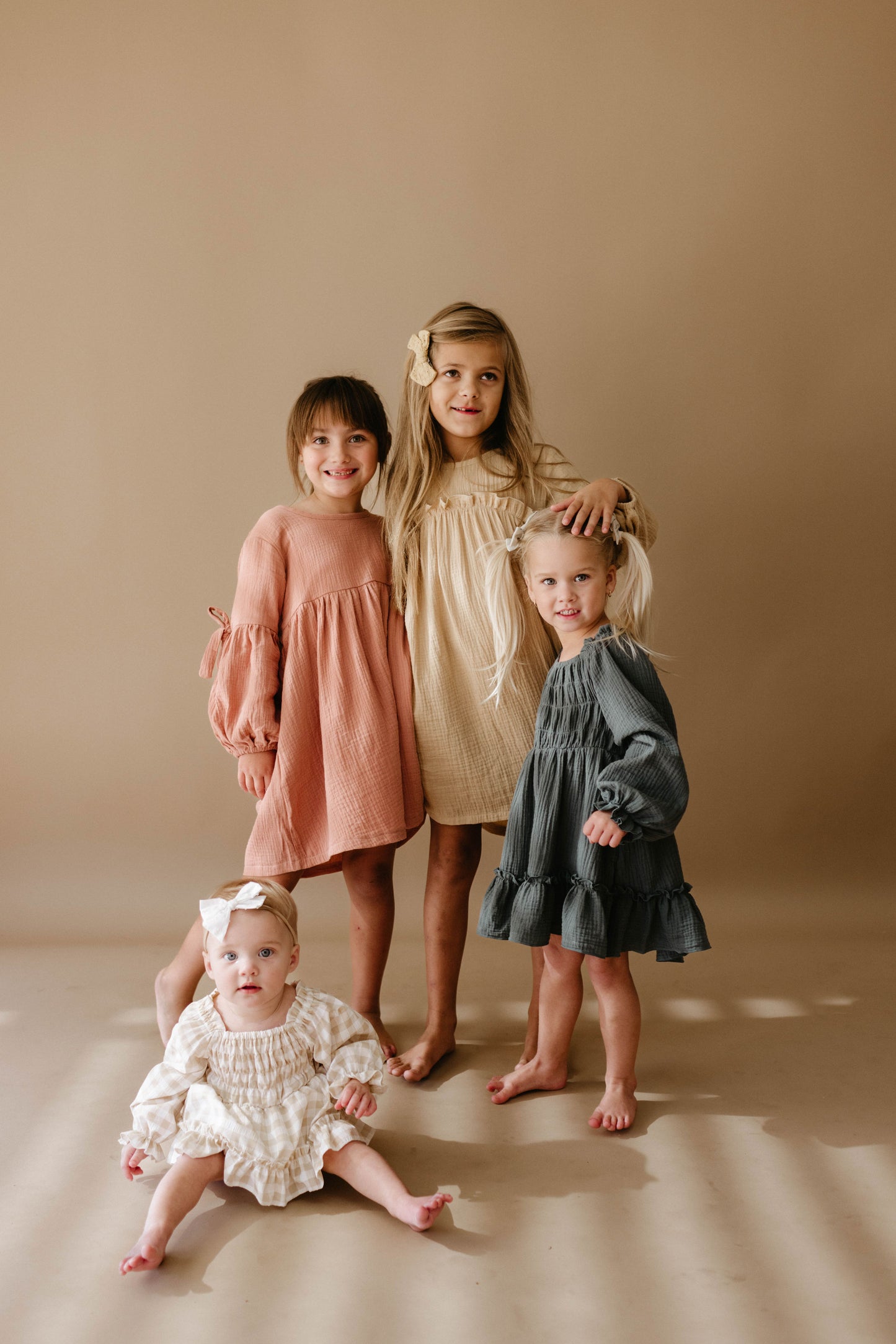 Four young girls model forever french baby’s Child's Dress | Meadow in neutral tones against a beige backdrop—three stand side by side, while the youngest sits in front, all facing the camera.