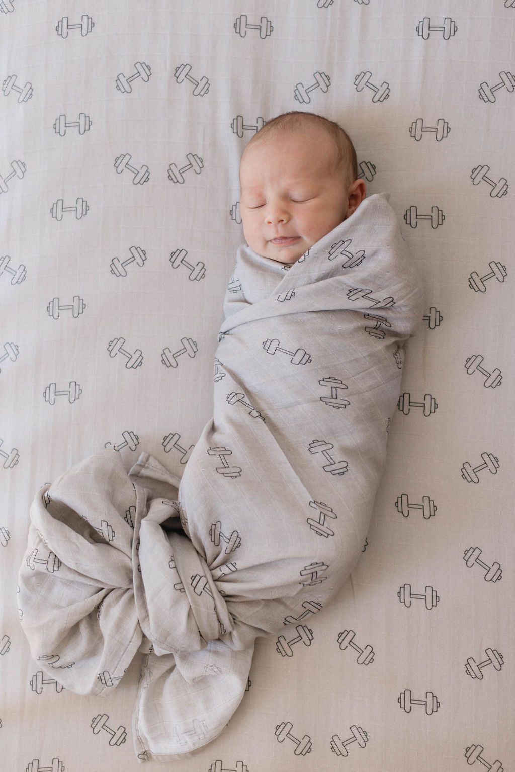 A newborn baby peacefully sleeps while swaddled in a 'Muslin Quilt | Arm Day + Black & Grey Checkerboard' from forever french baby, featuring an adorable dumbbell pattern. The baby is lying on a matching gray sheet, also decorated with dumbbells. Wrapped snugly, the quilt is tied off to one side, ensuring comfort and warmth.