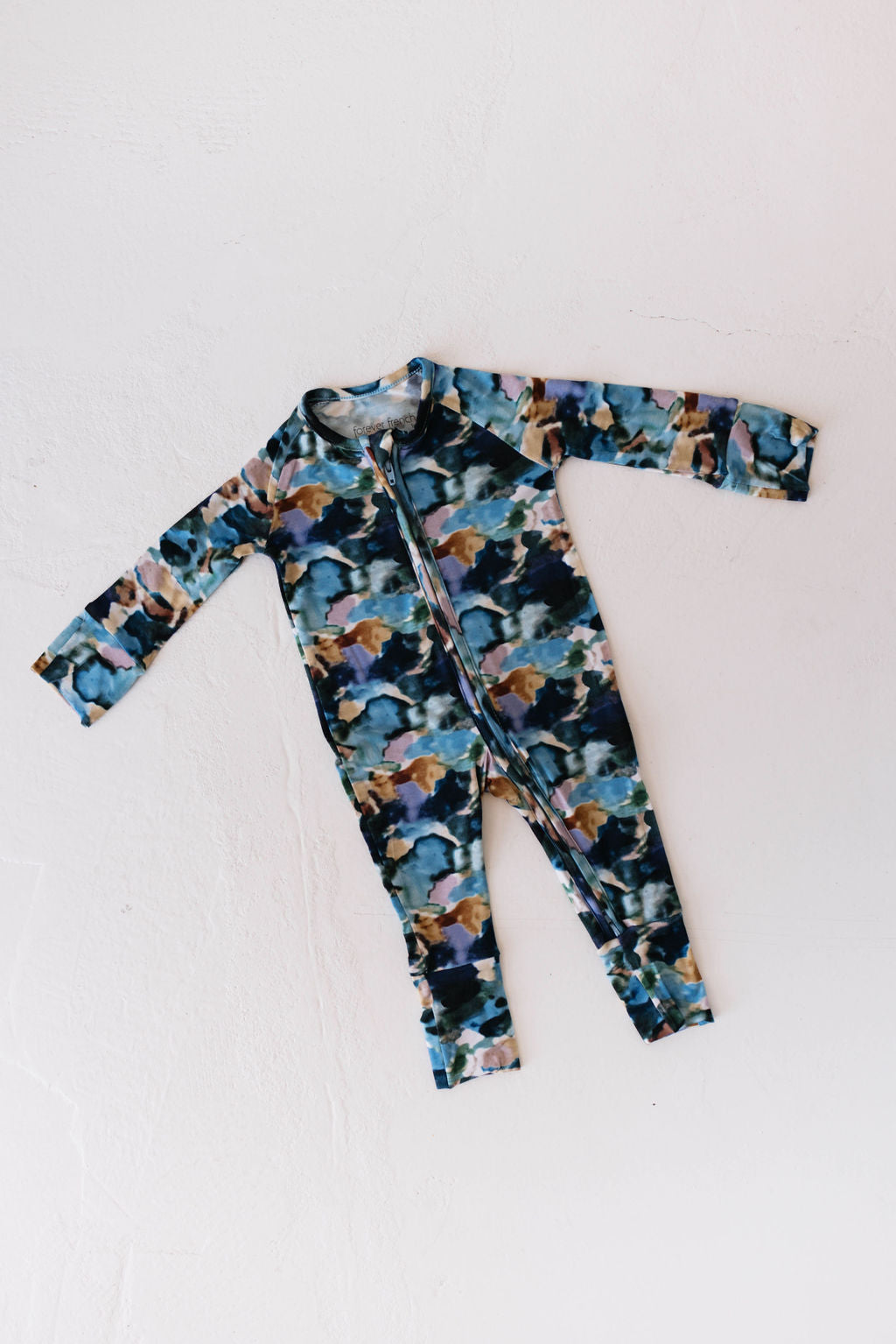 A full-length baby onesie with long sleeves and a front zipper lies flat on a white surface. These cozy "Bamboo Zip Pajamas | Charli Print" by forever french baby feature a colorful camouflage pattern in various shades of blue, green, brown, and beige.