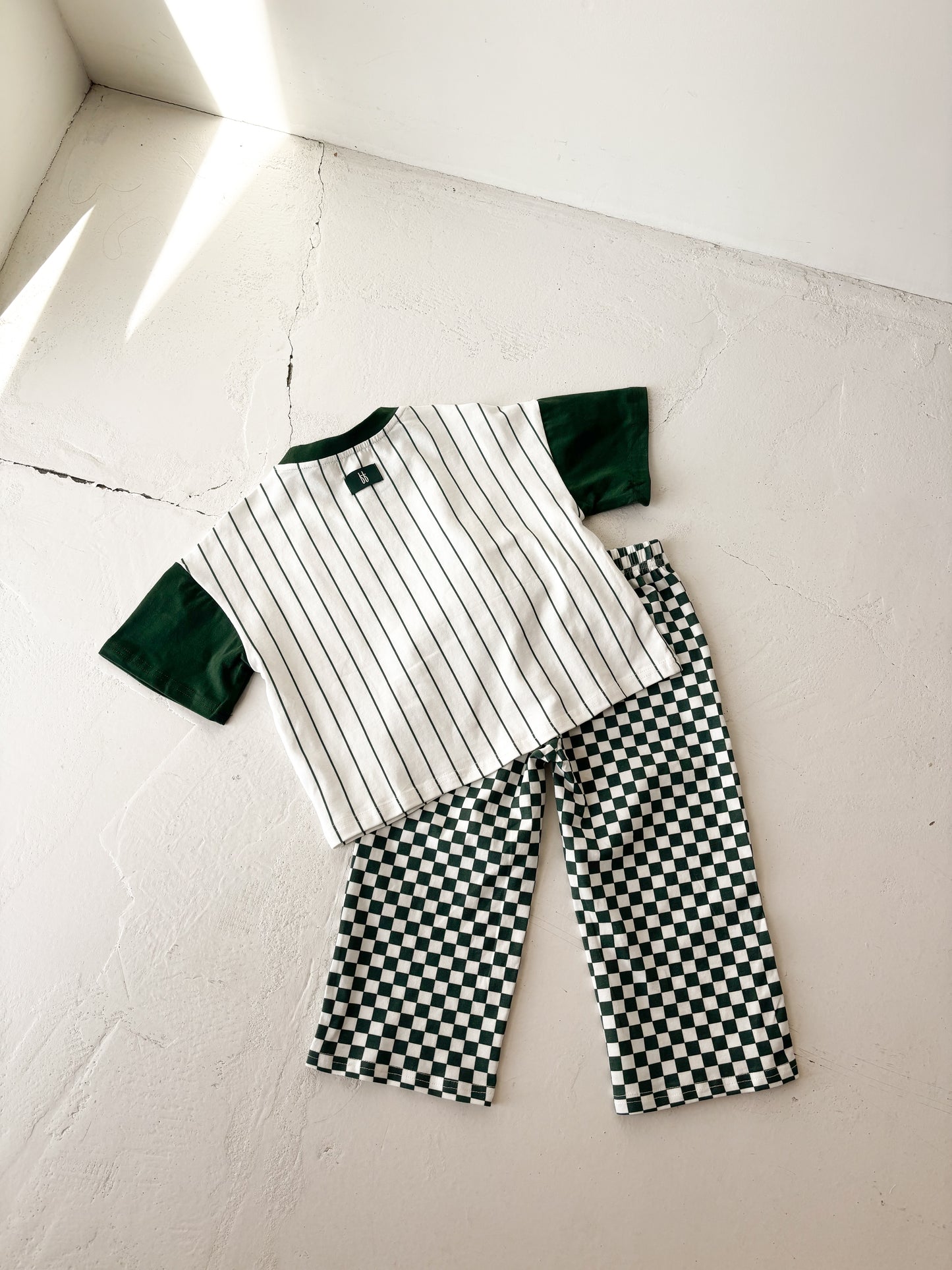 The forever french baby T-Shirt & Pant Set | Lucky Micro Check is displayed flat on a white surface, showing a striped short-sleeve shirt with green sleeves and green checkerboard wide leg pants. Soft sunlight creates gentle shadows nearby.