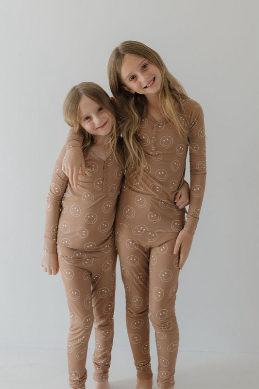 Two young girls smile in forever french baby's Bamboo Two Piece Pajamas | Sandstone Smile, featuring a white smiley face pattern. Their matching, breathable sleepwear keeps them comfy as they pose together against a light background.