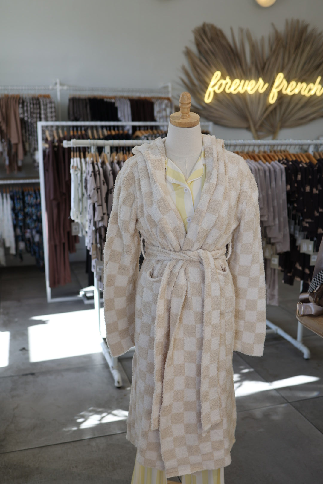 A mannequin wears the forever french baby Adult Robe in Cream Checkerboard, Oeko-Tex certified, in a boutique display enhanced by clothing racks and a neon "forever french" sign.