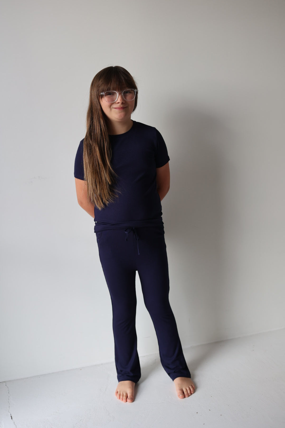 A young girl with long brown hair and glasses stands barefoot against a white wall, wearing the forever french baby Child Short Sleeve Bamboo Lounge Set in Blueberry Ribbed, smiling comfortably in these hypo-allergenic kids clothes.