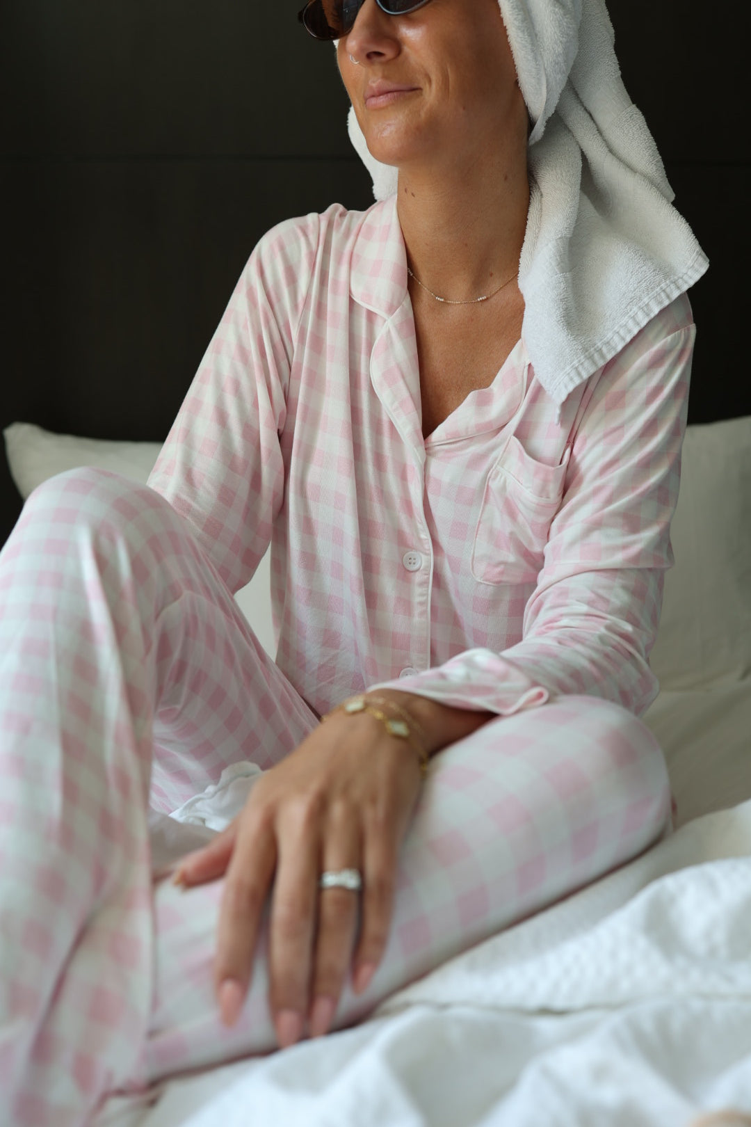 A woman in the forever french baby Long Sleeve Women's Bamboo Pajama in Pink Sugar sits on a bed, wearing a towel on her head and sunglasses, with bracelets and a ring. She smiles slightly.