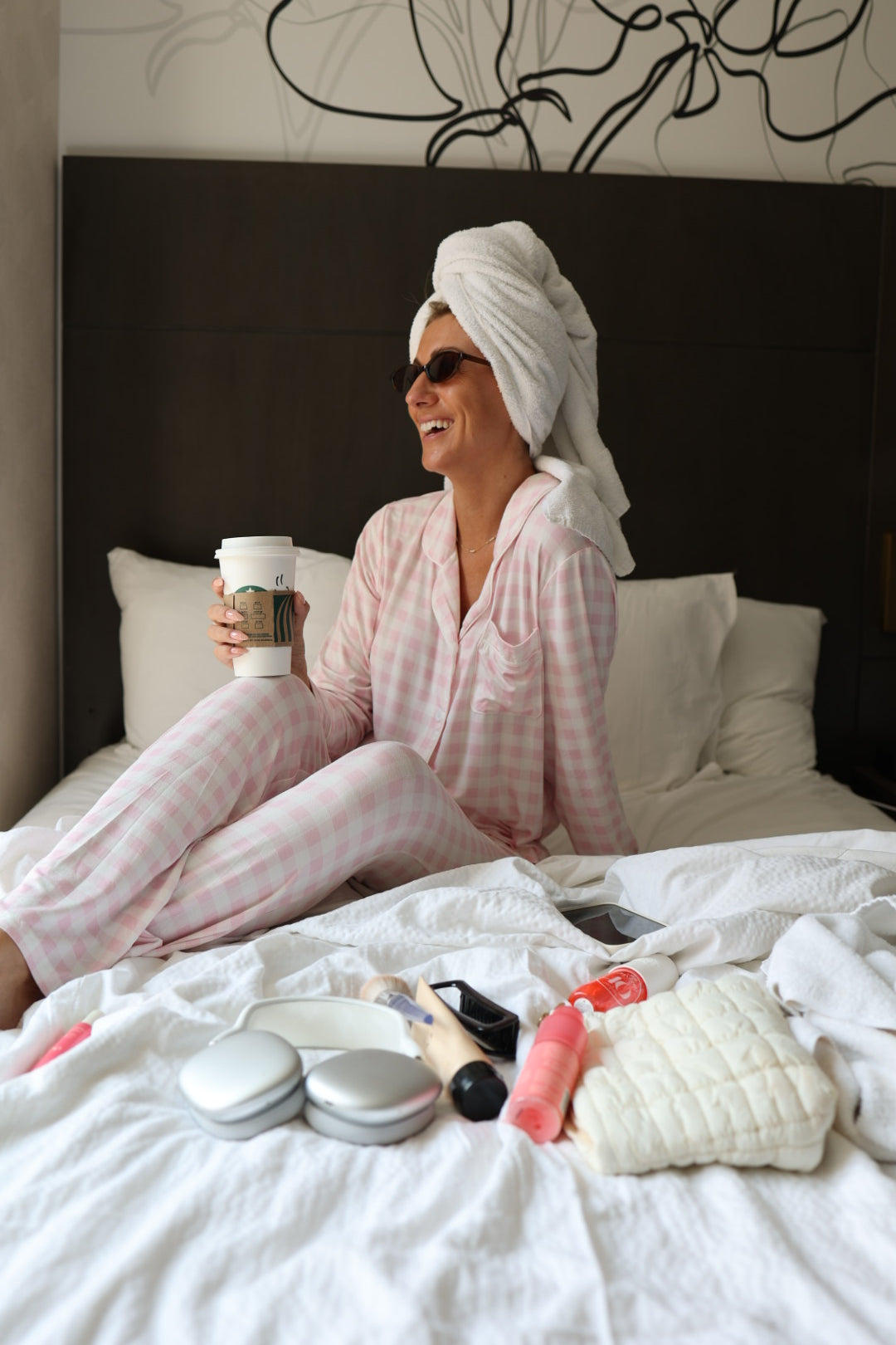 A woman wearing the forever french baby Long Sleeve Women's Bamboo Pajama in Pink Sugar with a towel on her head sits on a bed, smiling and holding a coffee cup. Various beauty products and a small white bag are laid out in front of her.
