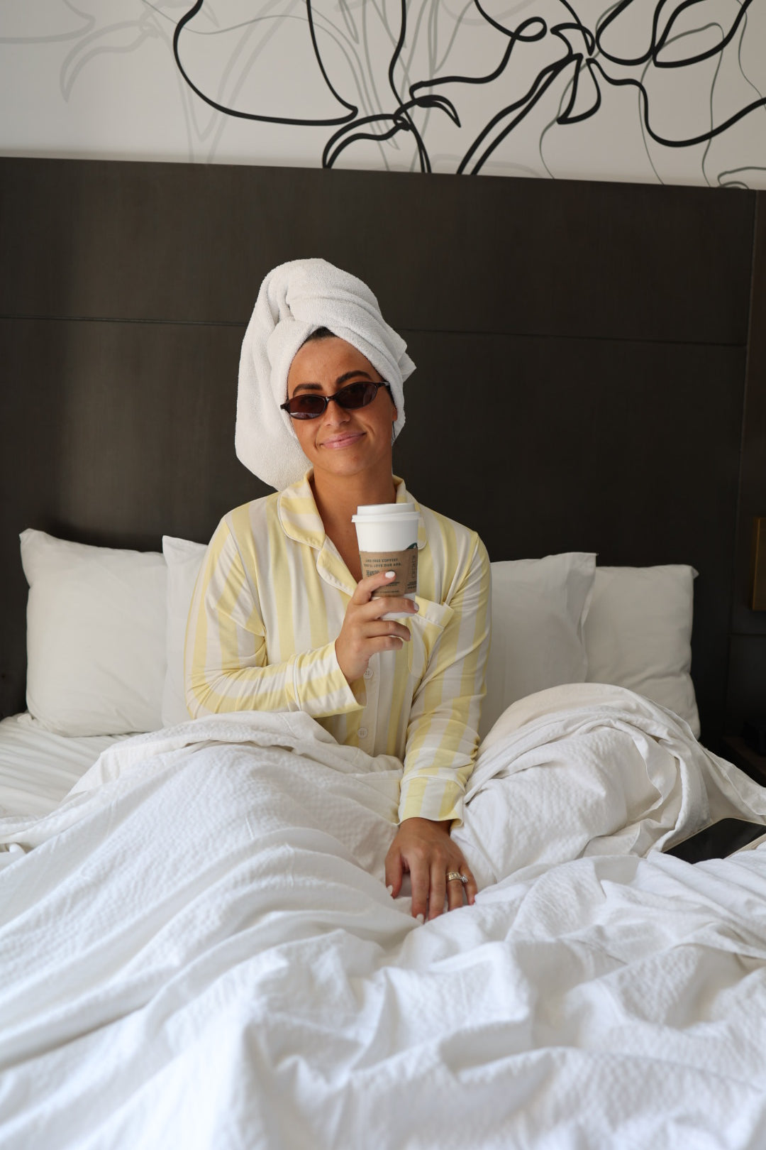 A woman in forever french baby’s Long Sleeve Women's Bamboo Pajama | Butter smiles in bed, wearing yellow-striped lounge pants and sunglasses, with a towel-wrapped head and takeaway coffee cup, surrounded by white bedding.