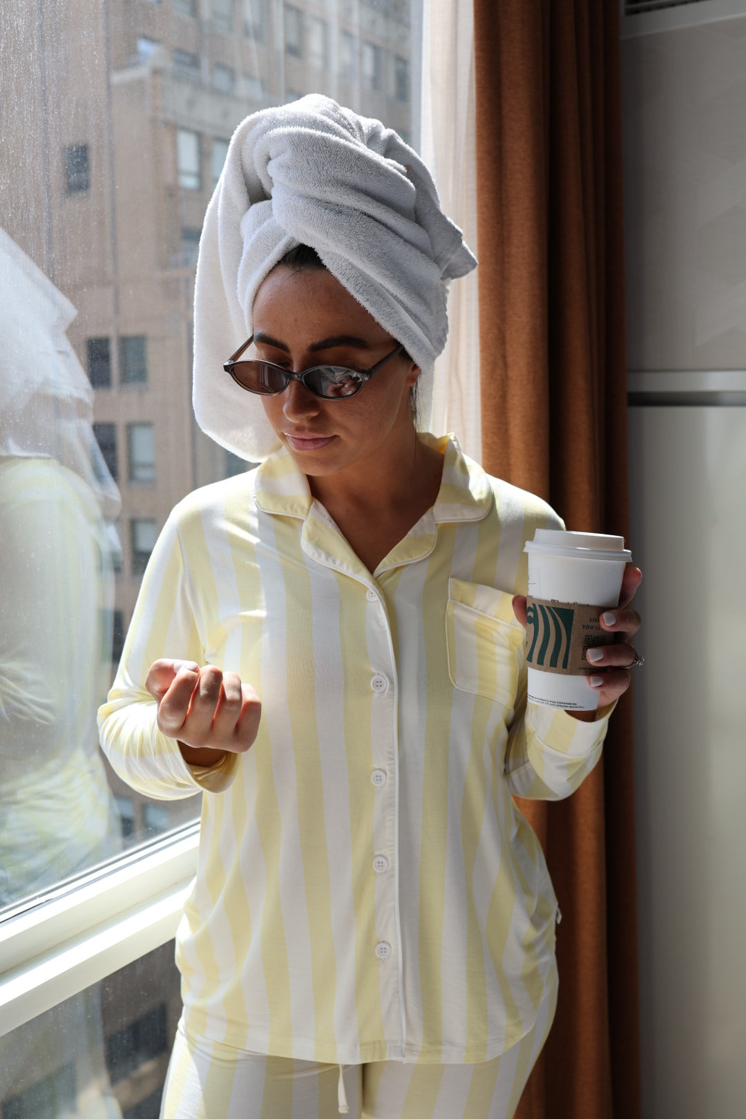Wearing forever french baby’s Long Sleeve Women's Bamboo Pajama in Butter, a woman with a towel wrapped around her head stands by a window, sipping coffee and checking her phone as city buildings appear outside.