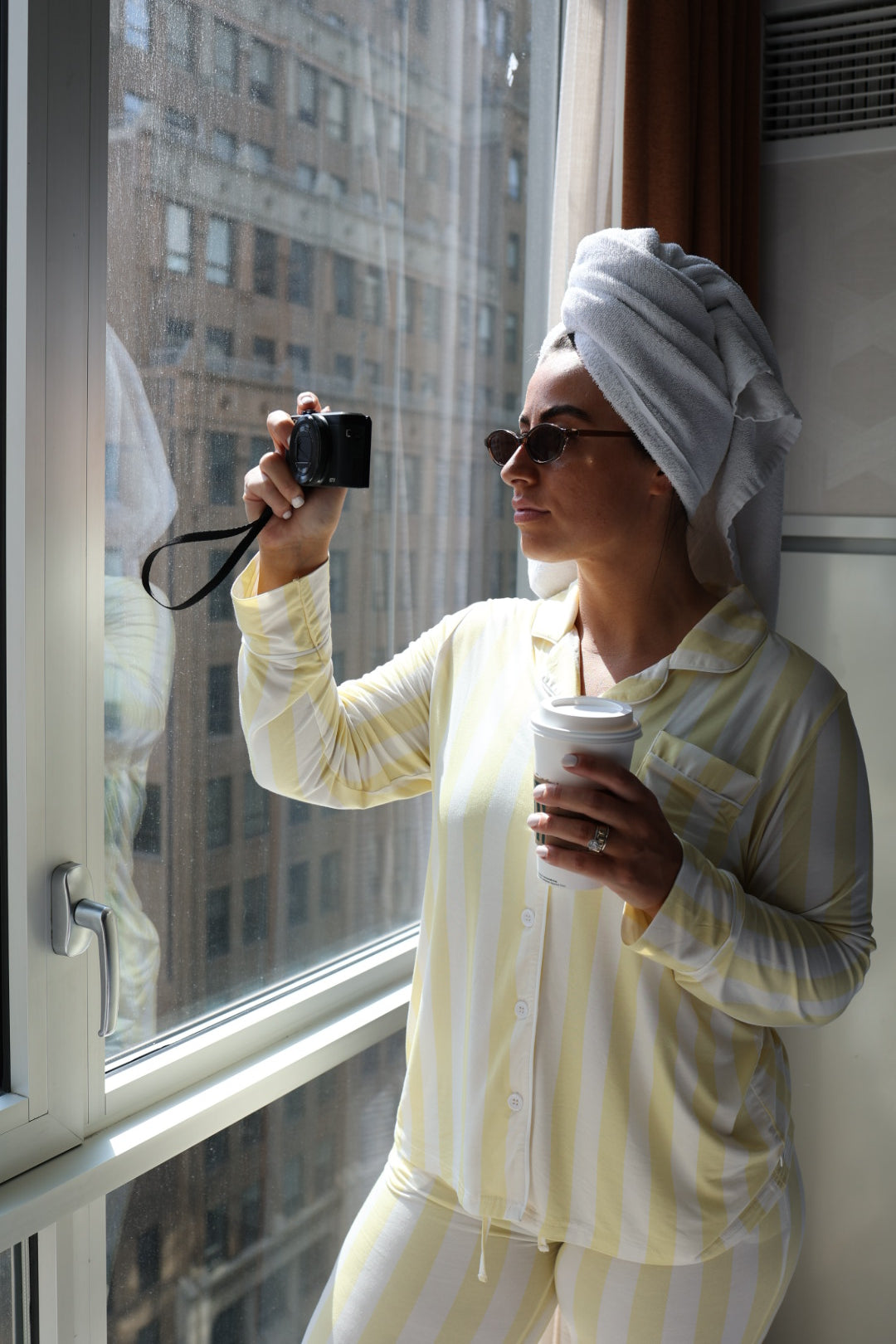 A woman in the Long Sleeve Women's Bamboo Pajama | Butter by forever french baby stands by a window with city views, wearing sunglasses and a towel on her head, holding a camera and coffee cup.