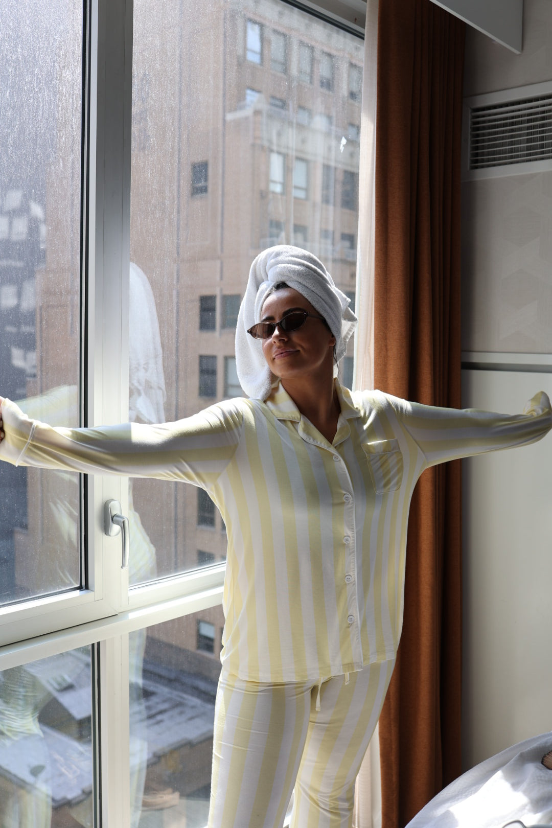 Wearing the Long Sleeve Women's Bamboo Pajama in Butter by forever french baby, a woman with sunglasses and a towel on her head stands by a window with arms outstretched as sunlight streams in, highlighting city buildings outside.