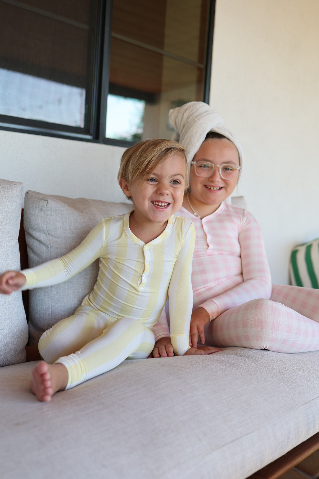 Two smiling children relax on a couch. The younger wears forever french baby Bamboo Two Piece Pajamas in Butter, while the older is in pink checkered hypo-allergenic sleepwear with a towel on their head. Both look happy indoors.