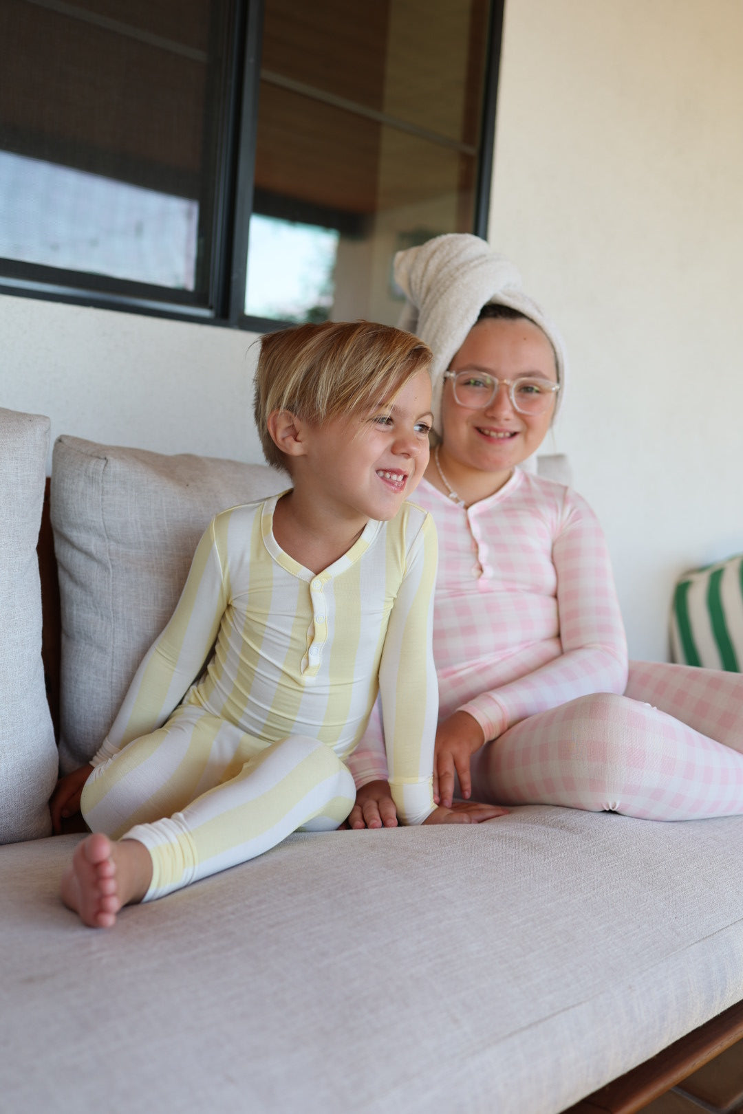 Two young children sit on a beige couch, smiling in their forever french baby Bamboo Two Piece Pajamas | Butter—one in yellow and white stripes, the other in pink checks with glasses and a towel, both enjoying their hypo-allergenic sleepwear.