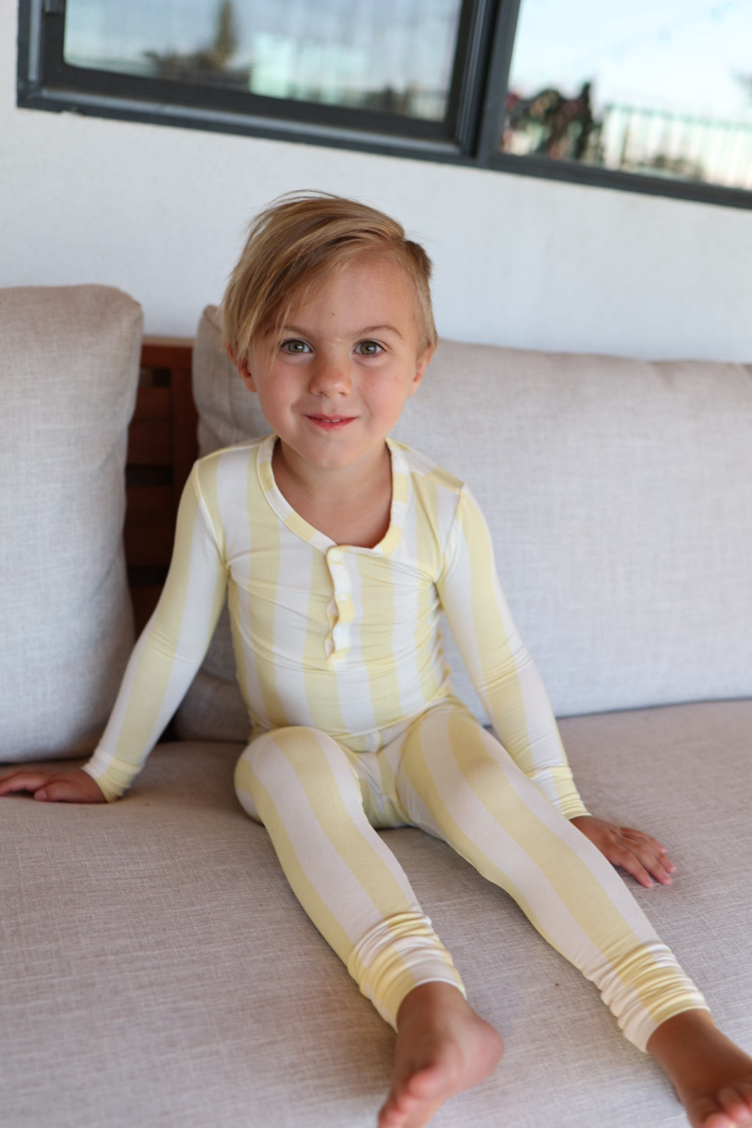 A young child sits barefoot on a light couch, smiling at the camera in cozy, hypo-allergenic forever french baby Bamboo Two Piece Pajamas in Butter.