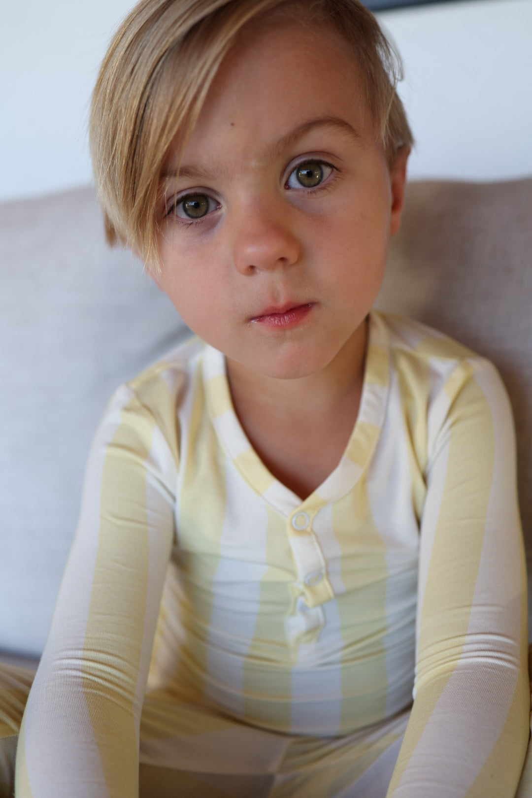 A young child with light brown hair and green eyes sits on a beige couch wearing forever french baby’s Bamboo Two Piece Pajamas in Butter, yellow and white striped hypo-allergenic sleepwear, looking at the camera with a neutral expression.