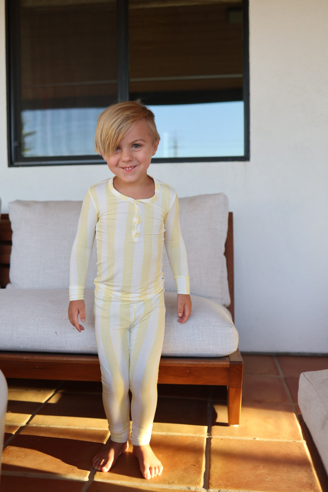 A young child with blond hair wears the forever french baby Bamboo Two Piece Pajamas in Butter, standing barefoot on a patio before an outdoor cushioned bench and smiling at the camera.