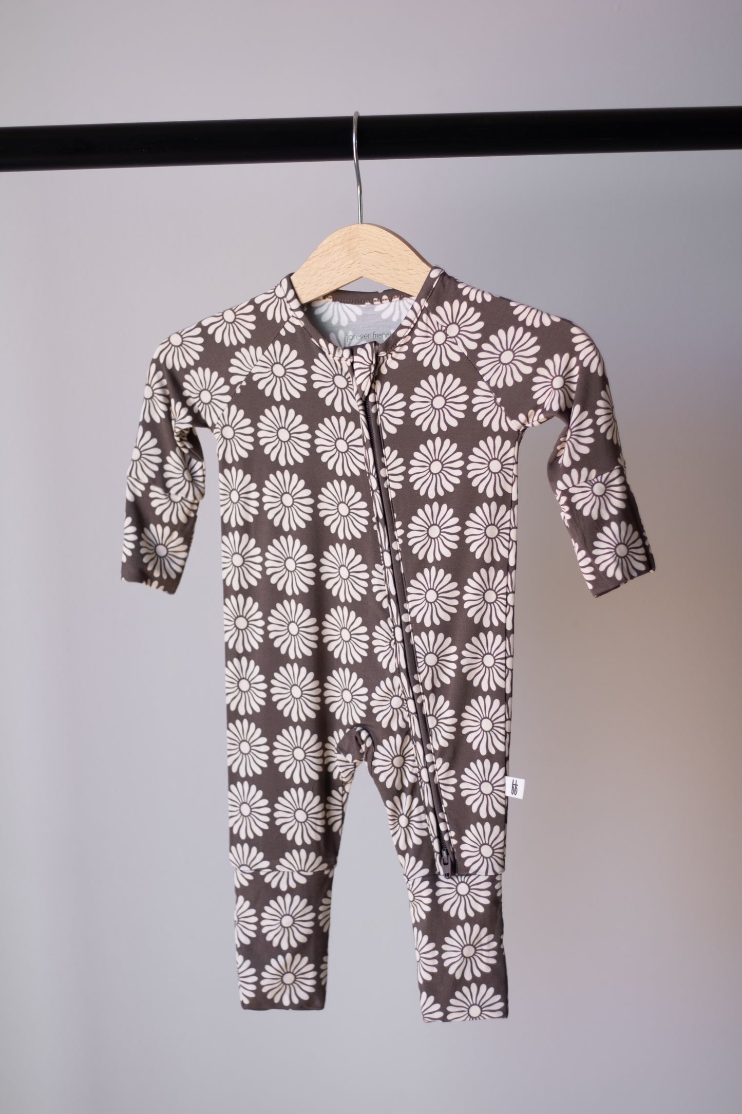 The Bamboo Zip Pajamas | Retro Floral by forever french baby, a brown long-sleeve, full-leg onesie with white daisy patterns, hangs on a wooden hanger against a light gray background—ideal breathable sleepwear for your baby.