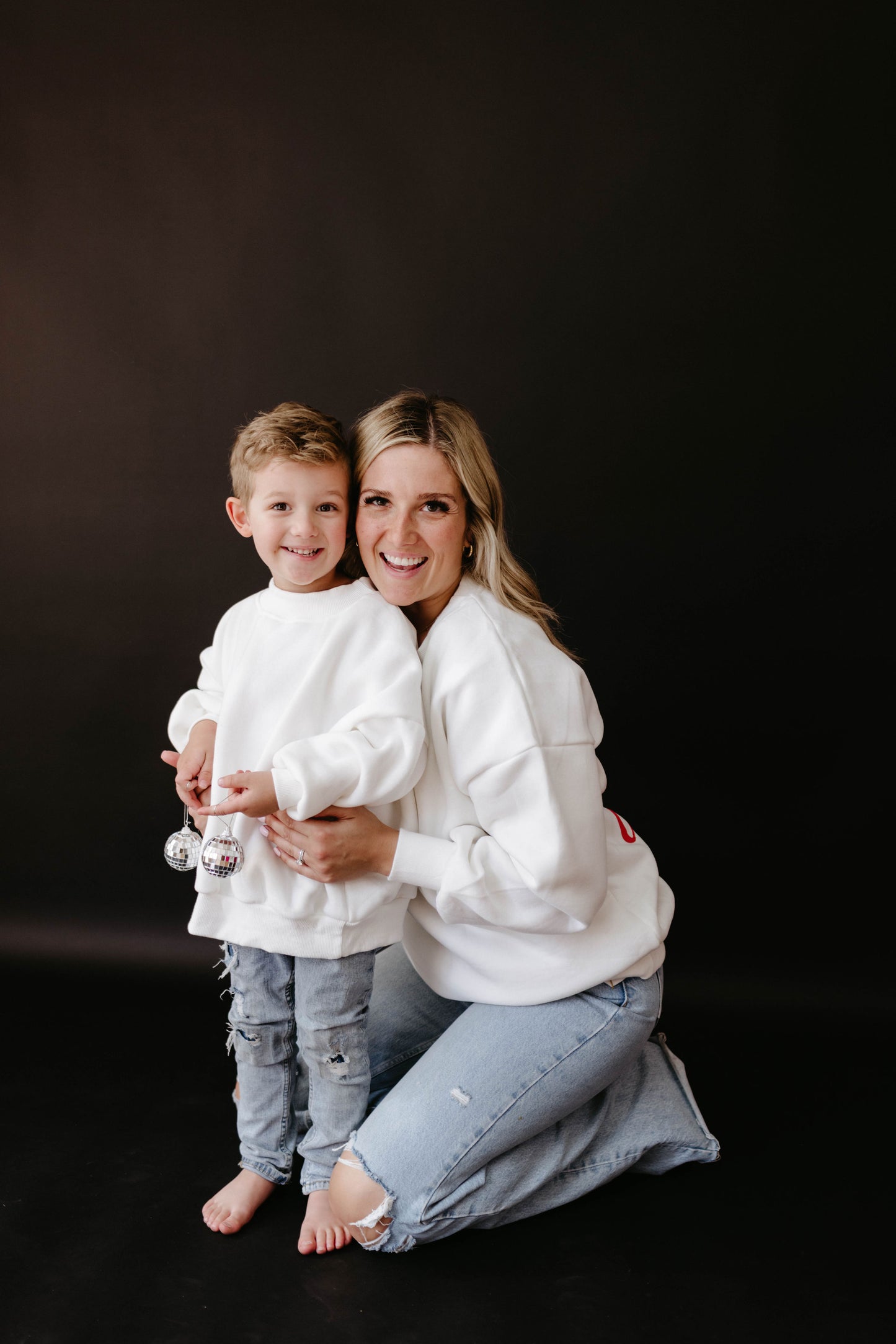 A smiling woman kneels beside a young child against a plain dark background. Both are wearing white "Baby It's Cold Outside" sweatshirts by forever french baby, perfect for the holiday season. The child excitedly holds two small ornaments as the woman lovingly wraps her arm around them, and they both look happy.