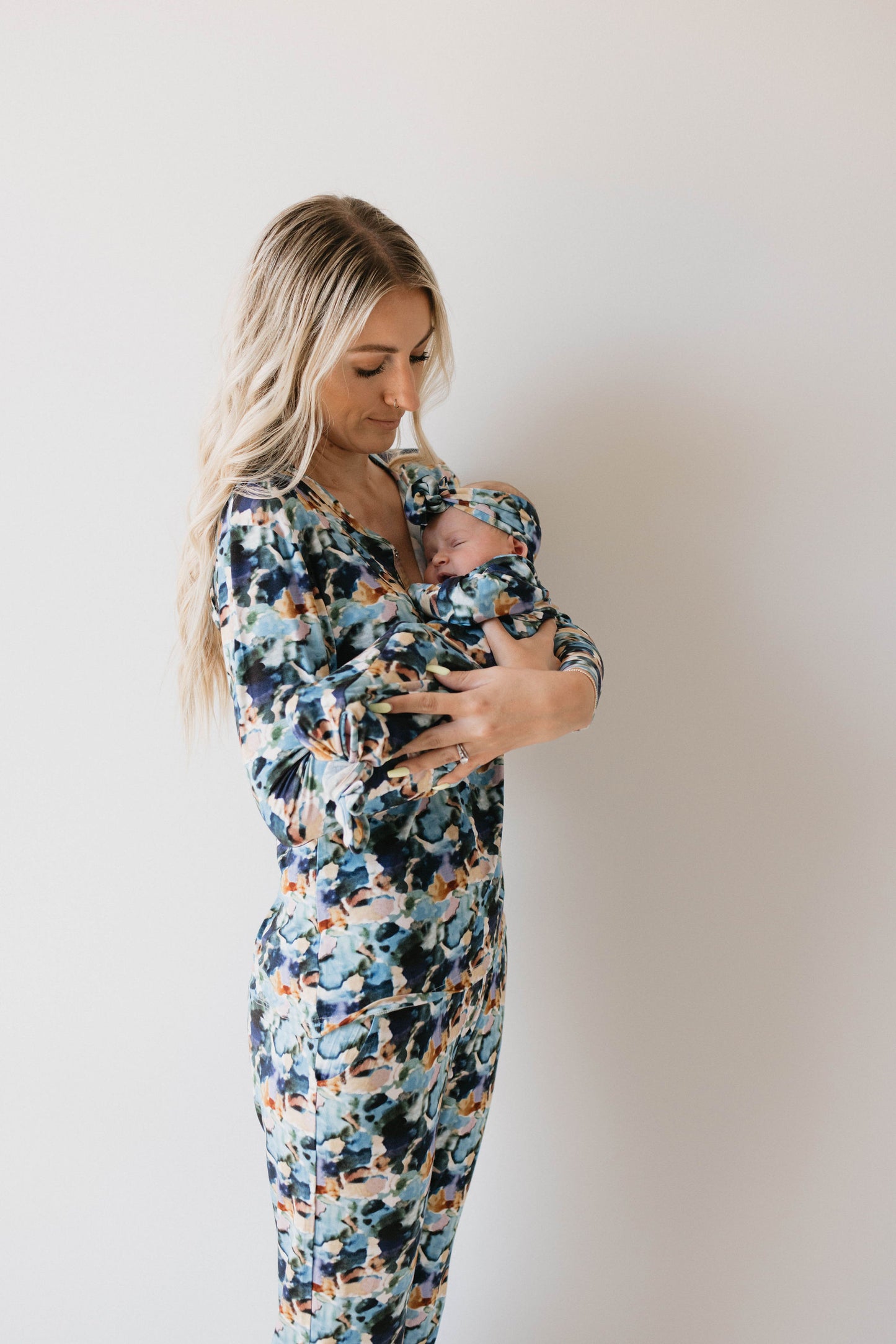 A woman with long, blonde hair wearing a matching floral patterned outfit is holding a newborn baby swaddled in the Charli Print Bamboo Knotted Gown by forever french baby. She is looking down at the baby lovingly against a plain, light-colored background.